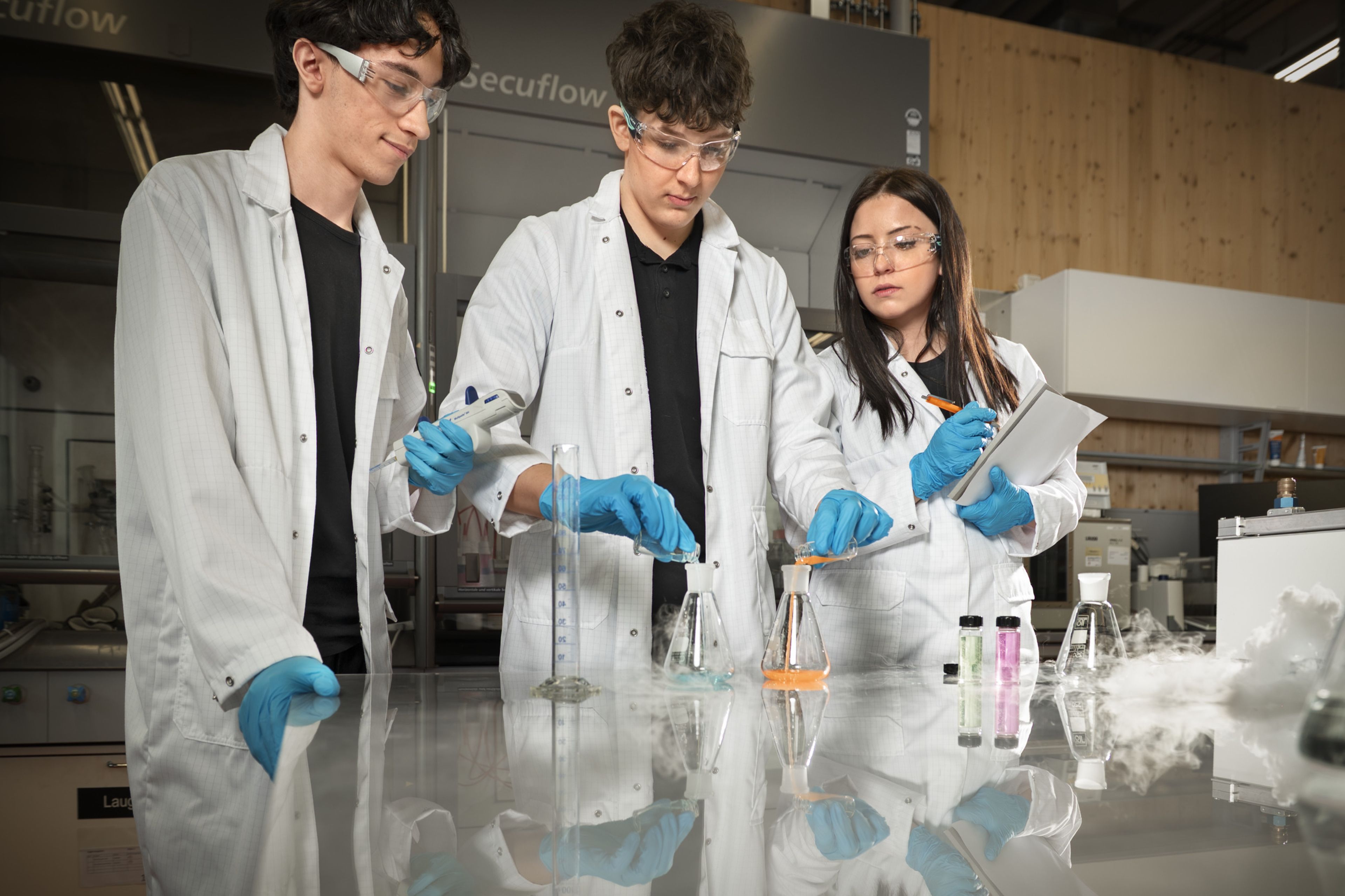 Auszubildende in der Ausbildung bei Blum im Beruf Kunststofftechnologie führen im Labor Versuche mit Kunststoffen durch, arbeiten mit Laborgeräten, Messflüssigkeiten und Schutzkleidung und dokumentieren die Ergebnisse gemeinsam.