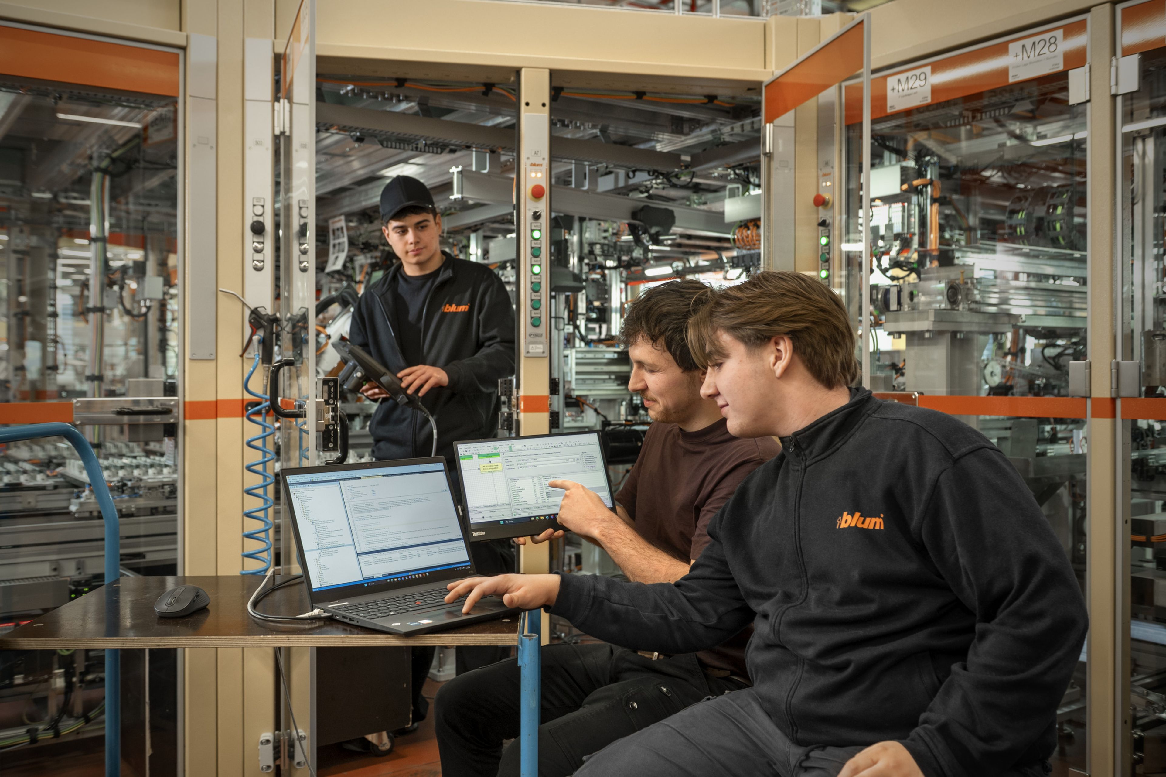 Mehrere Auszubildende bei Blum analysieren gemeinsam an einer laufenden Montageanlage mithilfe von Laptops und Diagnose-Software eine Störung, um Fehler in der automatisierten Anlage zu lokalisieren und zu beheben.