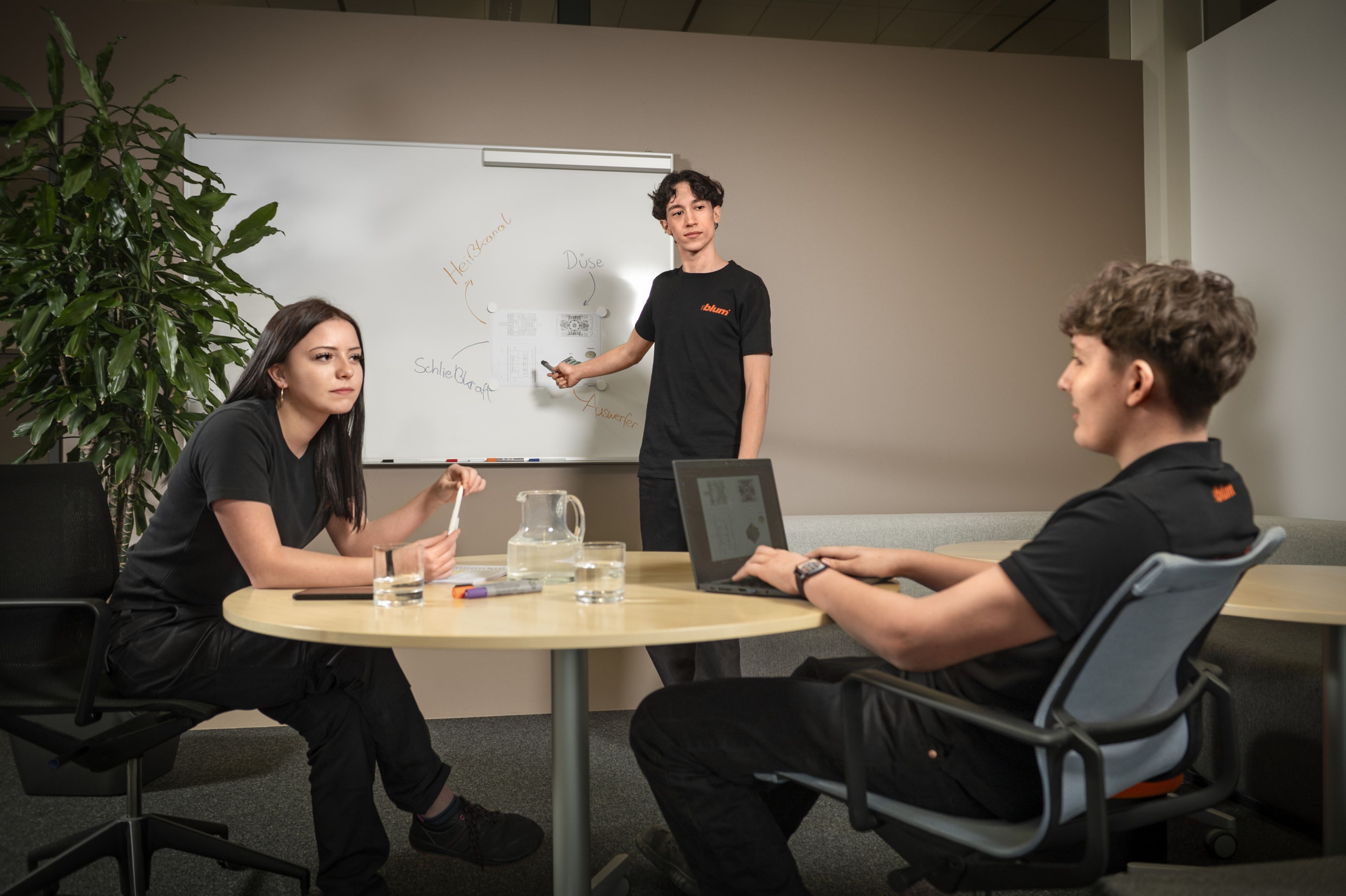 Auszubildende in der Ausbildung bei Blum im Beruf Kunststofftechnologie besprechen an einem Besprechungstisch Prozessabläufe, analysieren Zeichnungen und Skizzen am Whiteboard und arbeiten gemeinsam an der Planung technischer Lösungen.