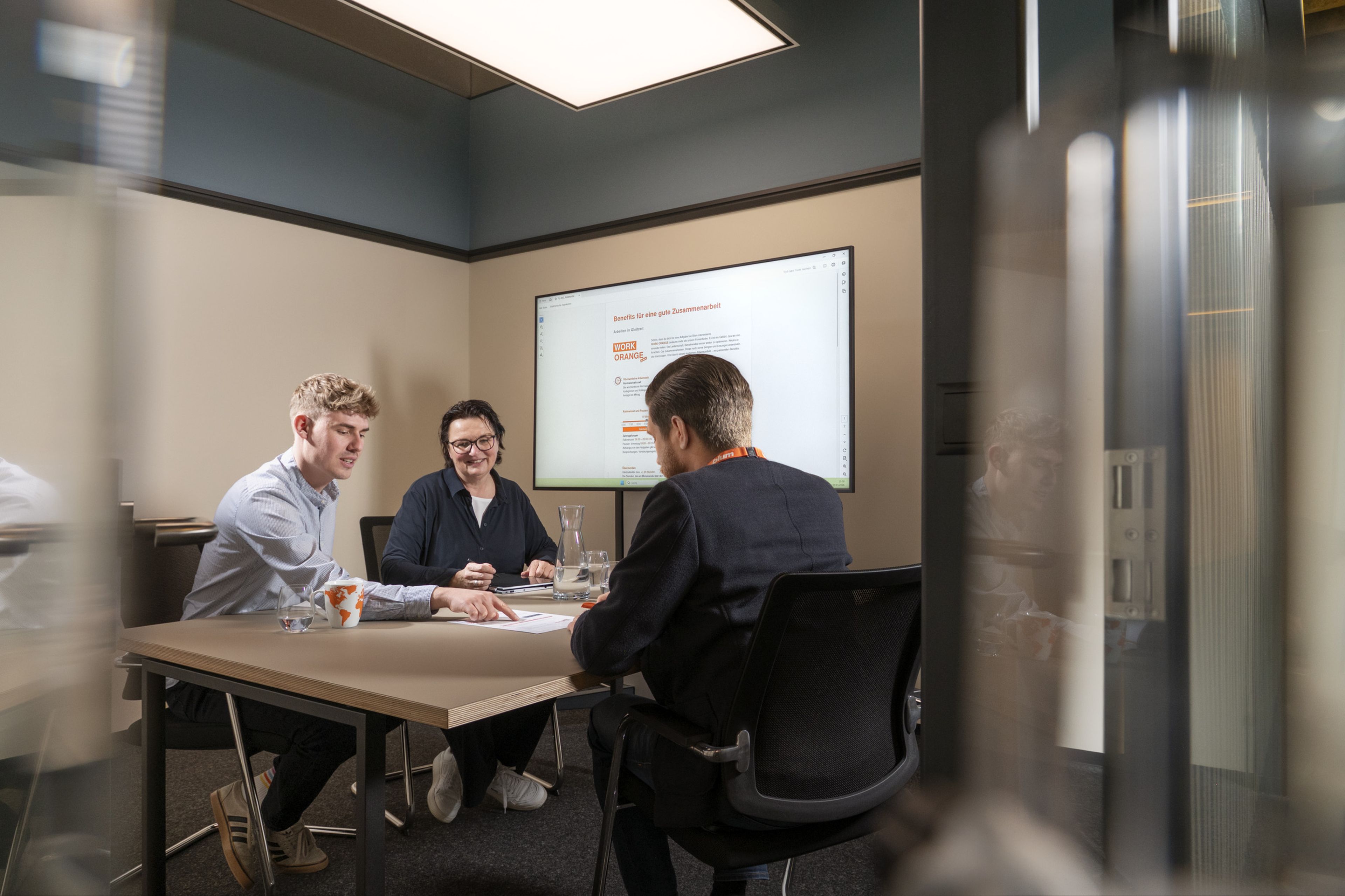 Three people in a meeting room, sitting at a table with a presentation displayed on a screen behind them.