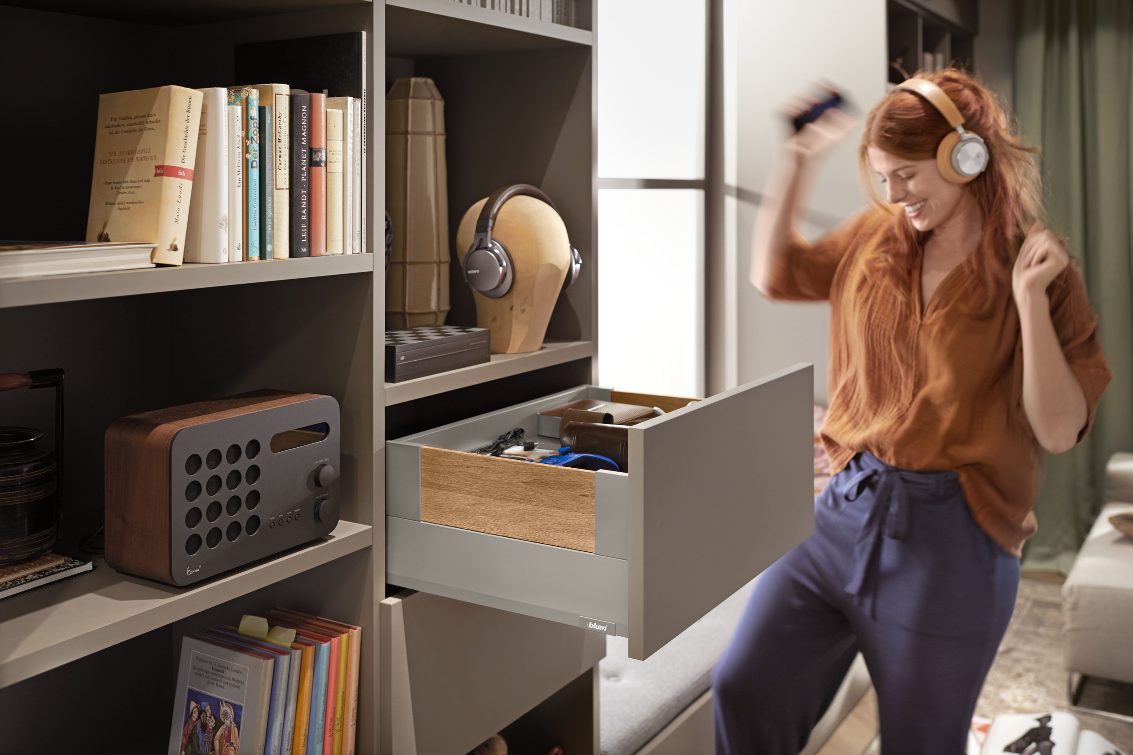 Eine Frau tanzt mit Kopfhörern auf dem Kopf, in dem Regal vor ihr ist eine Schublade geöffnet