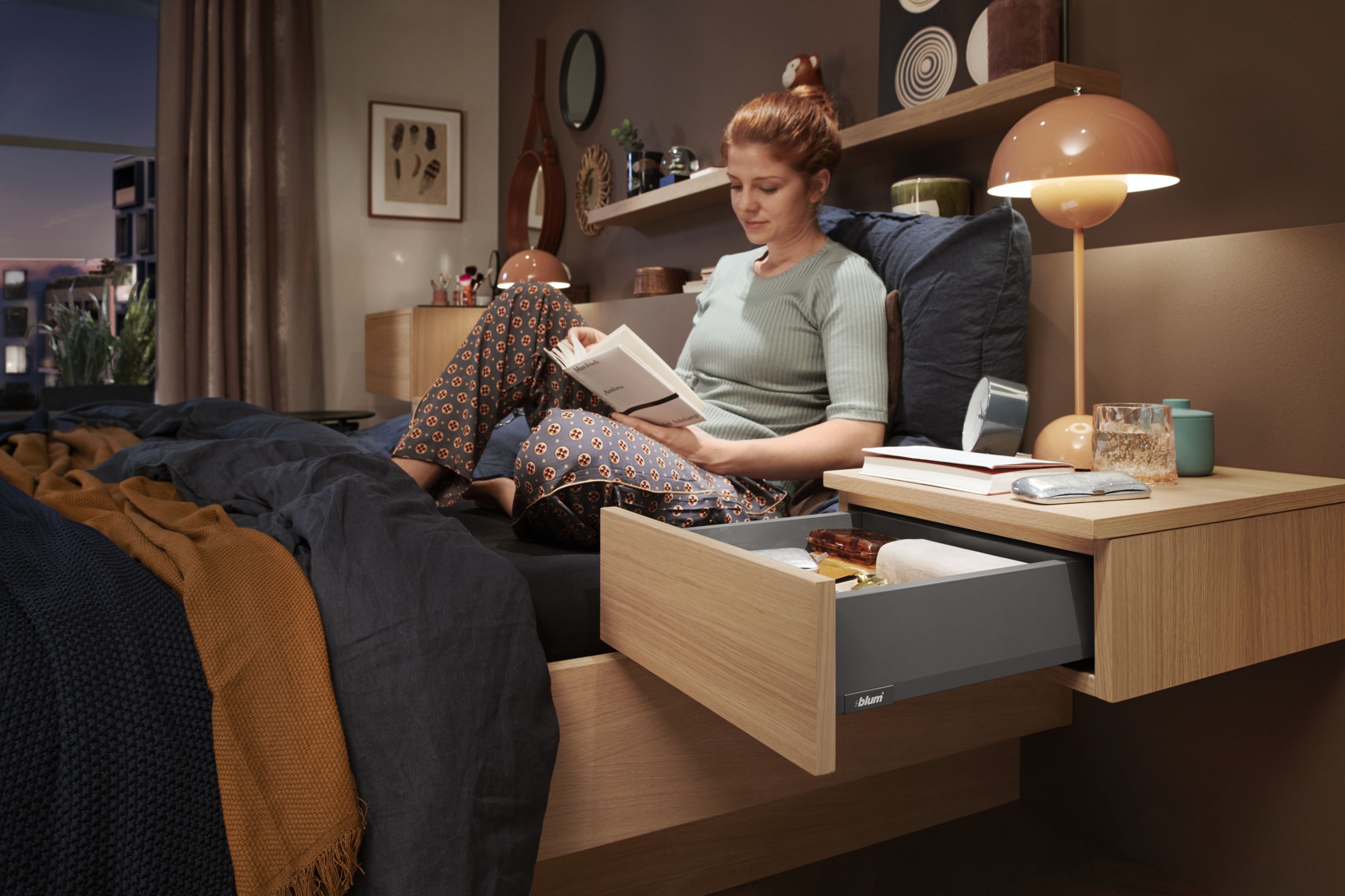 Eine Frau sitzt auf einem Bett und liest ein Buch, im Nachtkästchen neben ihr steht eine MERIVOBOX-Schublade offen