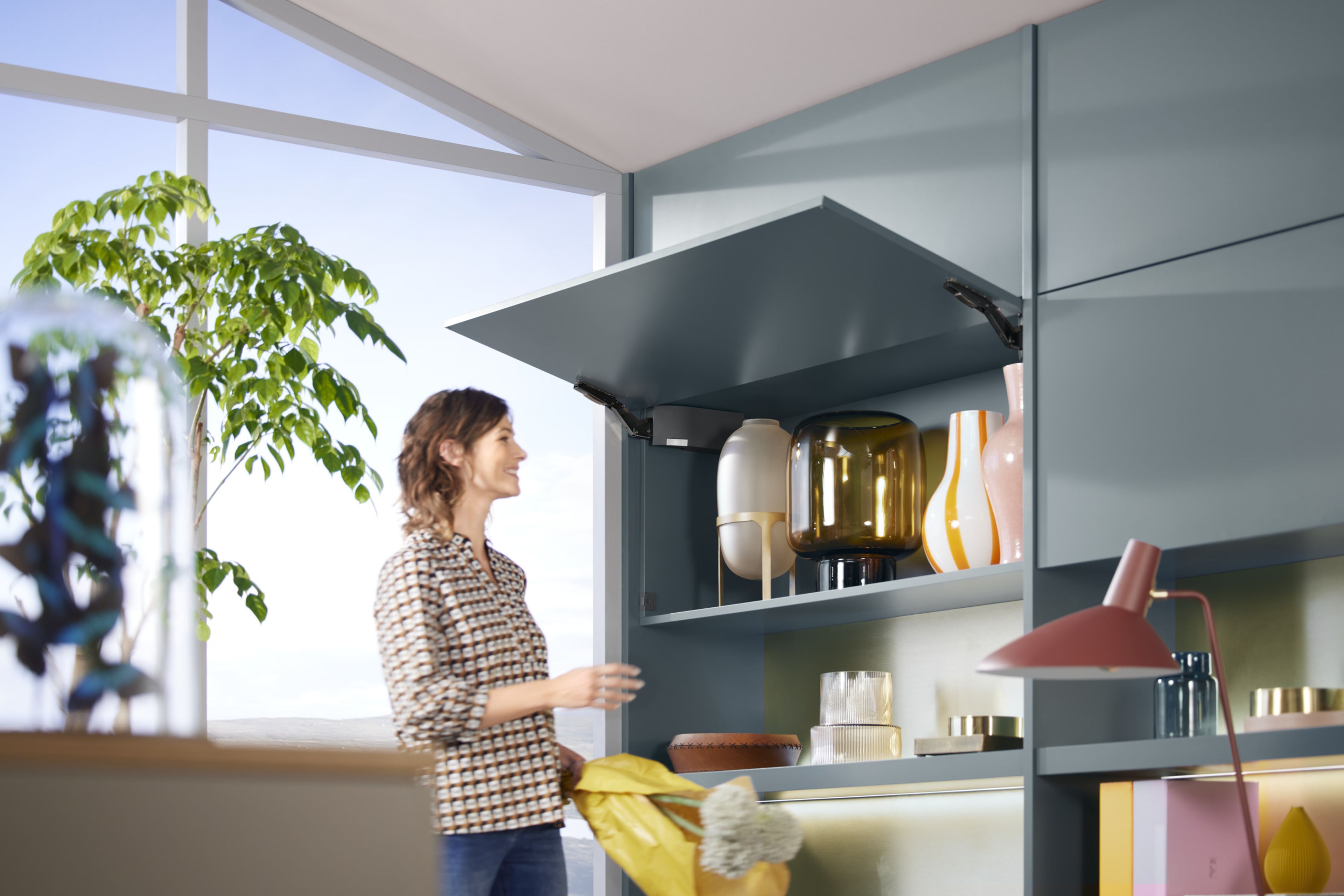 A woman stands in front of an open blue-green wardrobe with AVENTOS HK top
