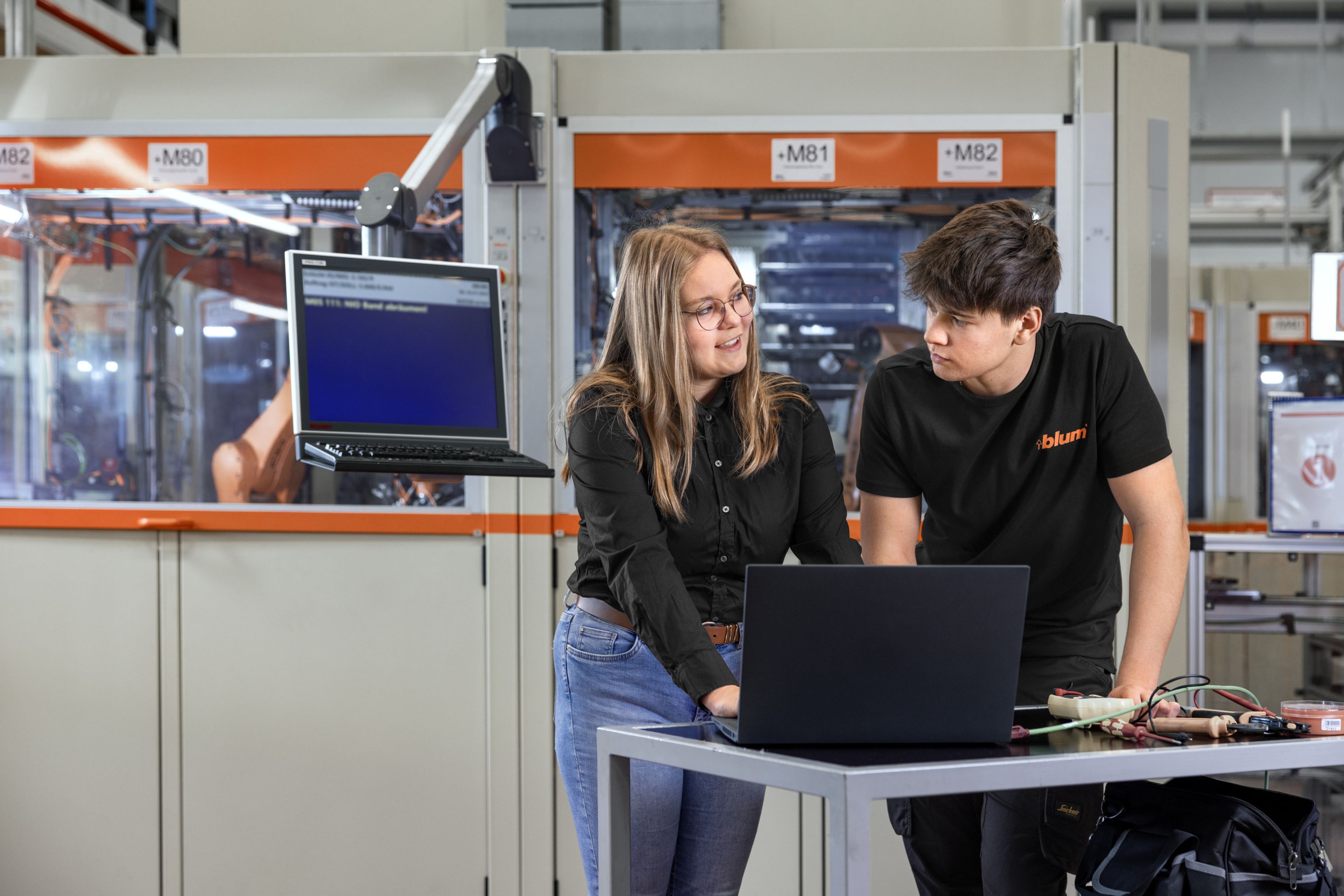 Das Bild zeigt einen Lehrling der gemeinsam mit seiner Ausbilderin an einer Hightech-Anlage eine Fehlerbehebung in der Programmierung durchführt. 