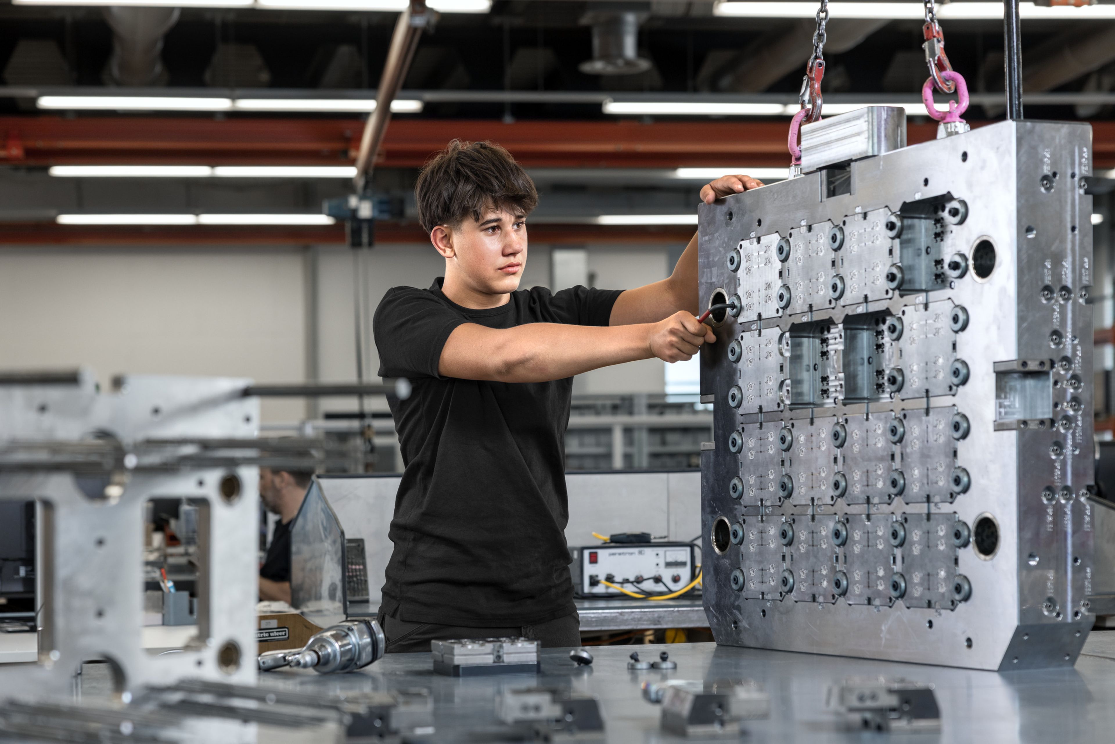 Auszubildender in der Mechanik bei Blum arbeitet in einer modernen Werkstatt an einem großen Metall-Werkzeugblock. Sichtbar sind präzise gefertigte Formeinsätze, Schraubwerkzeuge und weitere mechanische Komponenten auf dem Arbeitstisch. Im Hintergrund industrielle Umgebung mit Maschinen und Montageeinrichtungen.