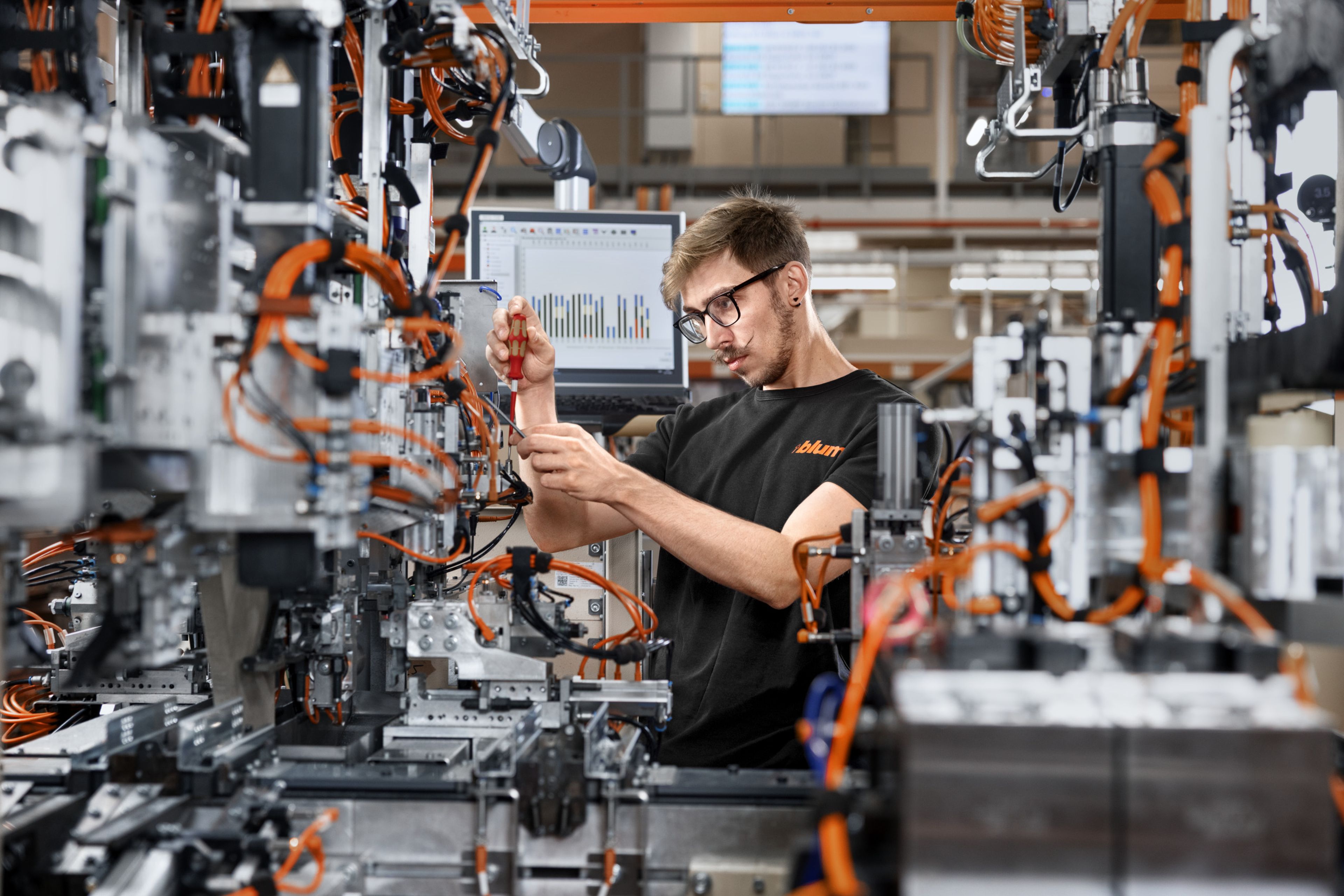 Auszubildende Person arbeitet in einer modernen Hightech‑Produktionsanlage an komplexen Maschinen und Kabelsystemen, während auf einem Monitor technische Diagramme angezeigt werden; die Szene veranschaulicht den praktischen Alltag einer Hightech‑Ausbildung in der Dualen Akademie nach einem beruflichen Neustart von theoretischem Studium hin zu angewandter Technologie.