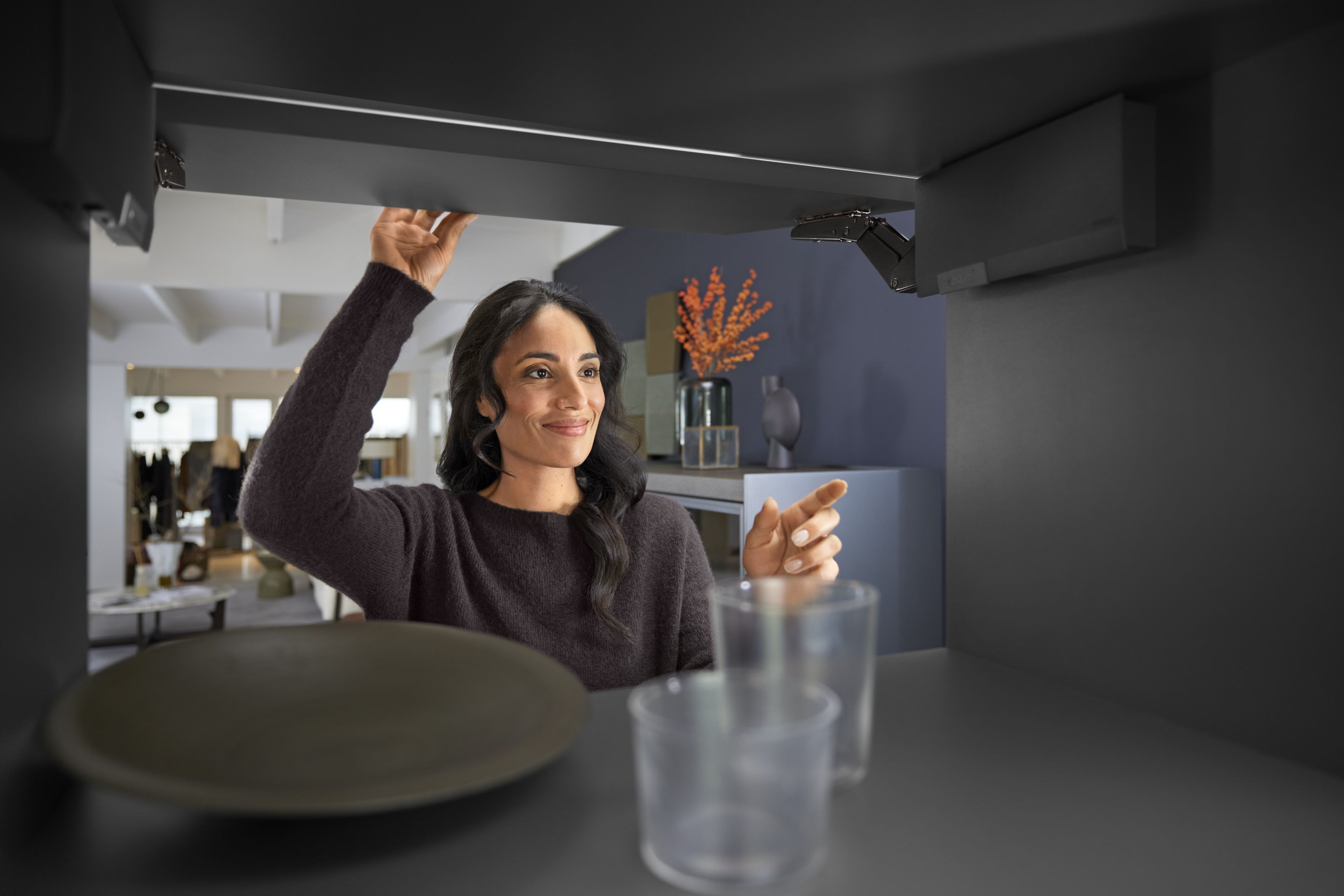 A woman opens a wall cabinet equipped with the AVENTOS HK top lift system with fittings in eclipse black