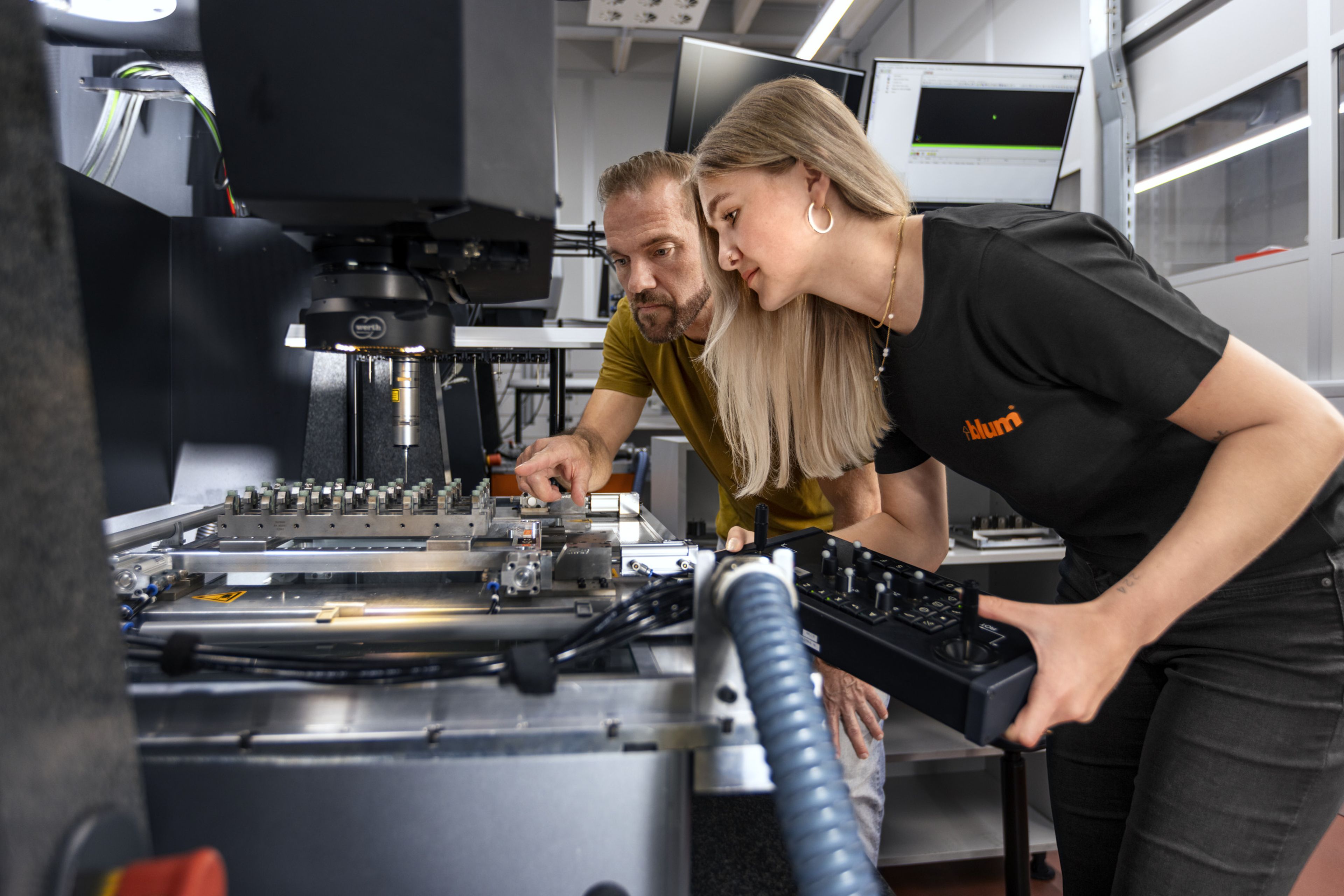 Das Bild zeigt einen Lehrling der Fertigungsmesstechnik, wie sie gemeinsam mit ihrem Ausbilder ein Bauteil mit einer Hightech-Maschine antastet für die Ausmessung. 