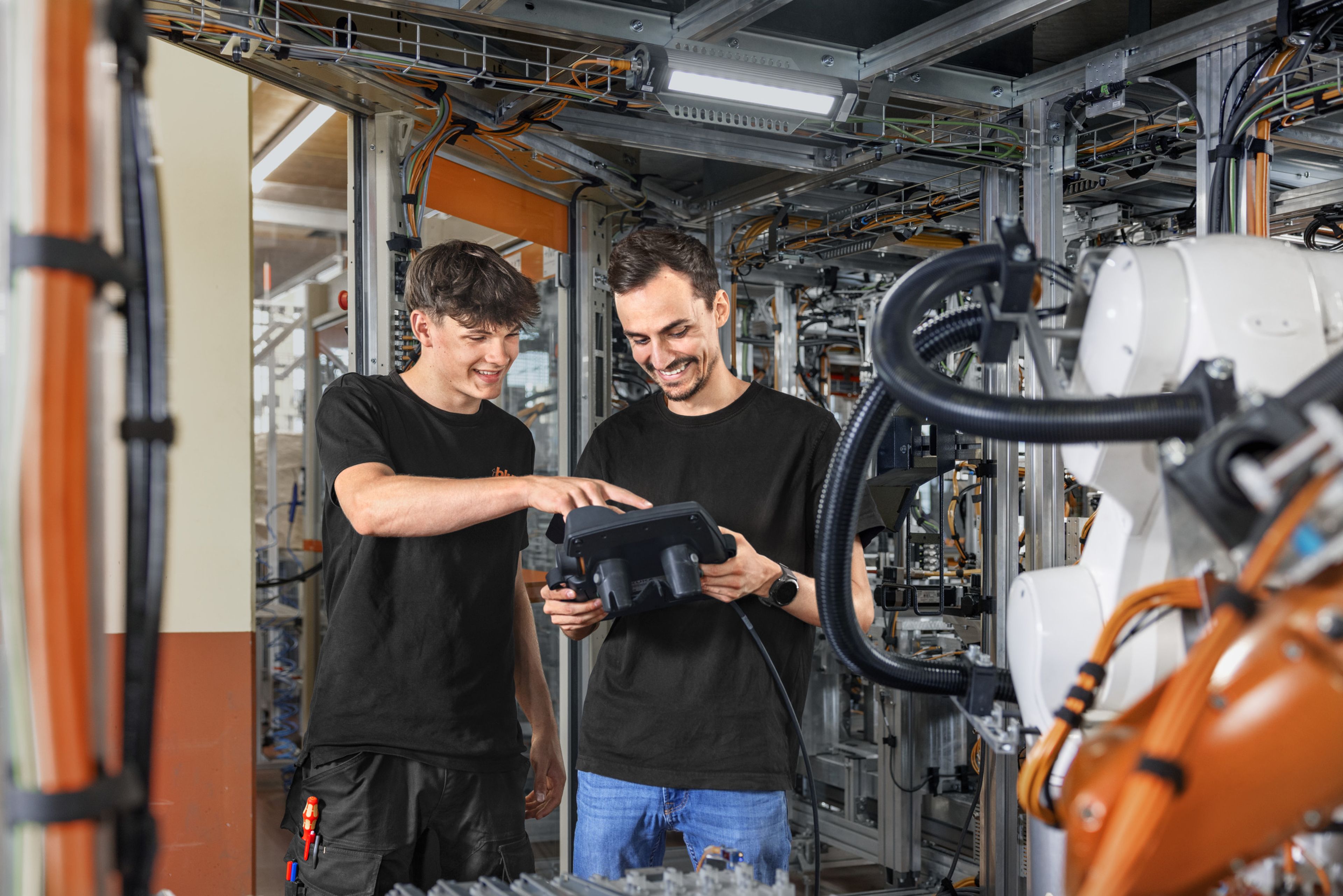 Das Bild zeigt einen Lehrling mit seinem Ausbilder wie sie sich über die Robotersteuerung in einer Hightech-Anlage unterhalten. 