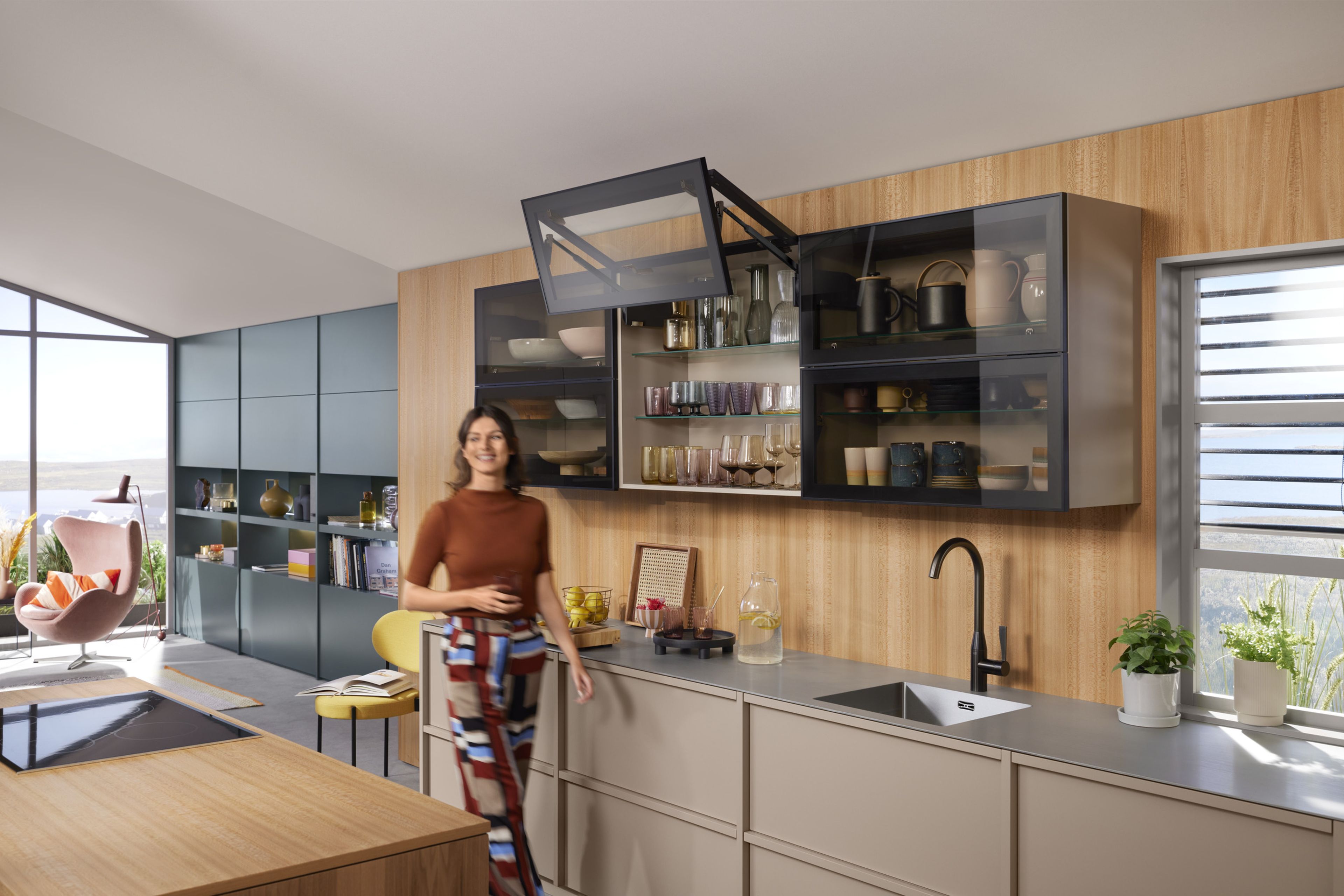 A woman stands in front of an open glass front with a black aluminium frame, equipped with the AVENTOS HF top bi-fold lift system