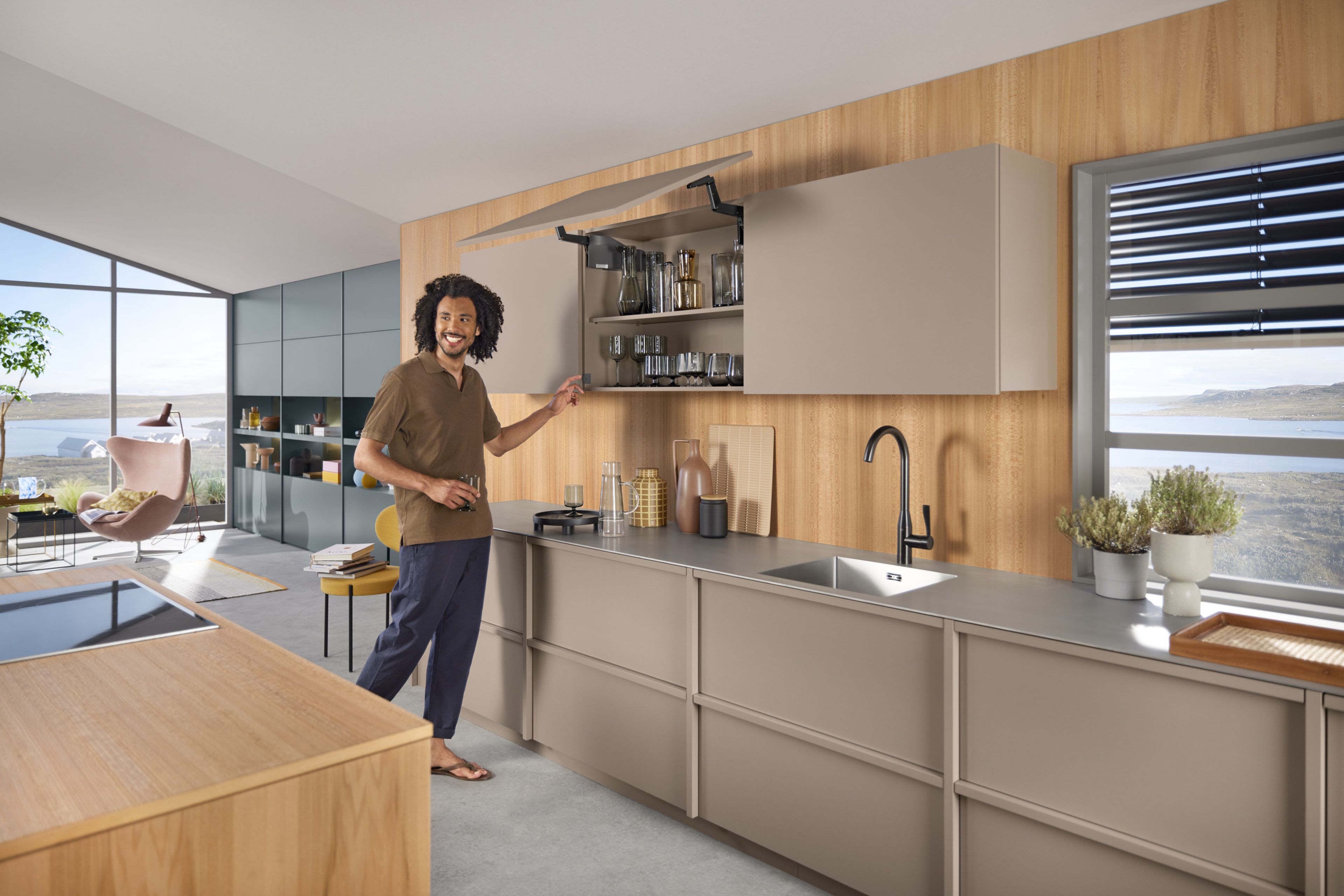 A man stands in a beige island kitchen with an open front fitted with an AVENTOS HS top up & over lift system