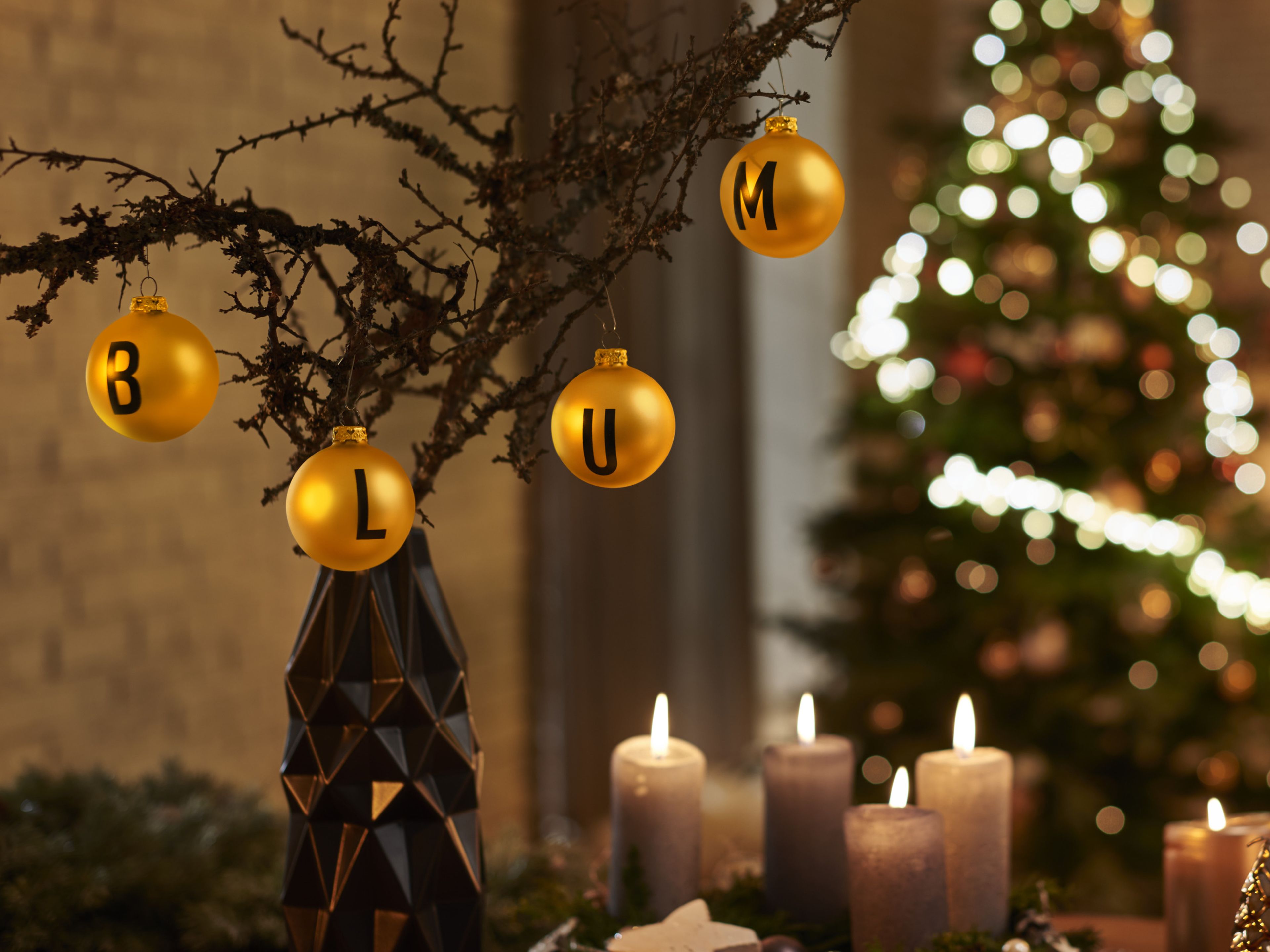 Goldfarbene Ornamente, die den Schriftzug „BLUM“ bilden, auf einem Ast in einer Vase, mit brennenden Kerzen und einem verschwommenen Weihnachtsbaum im Hintergrund.