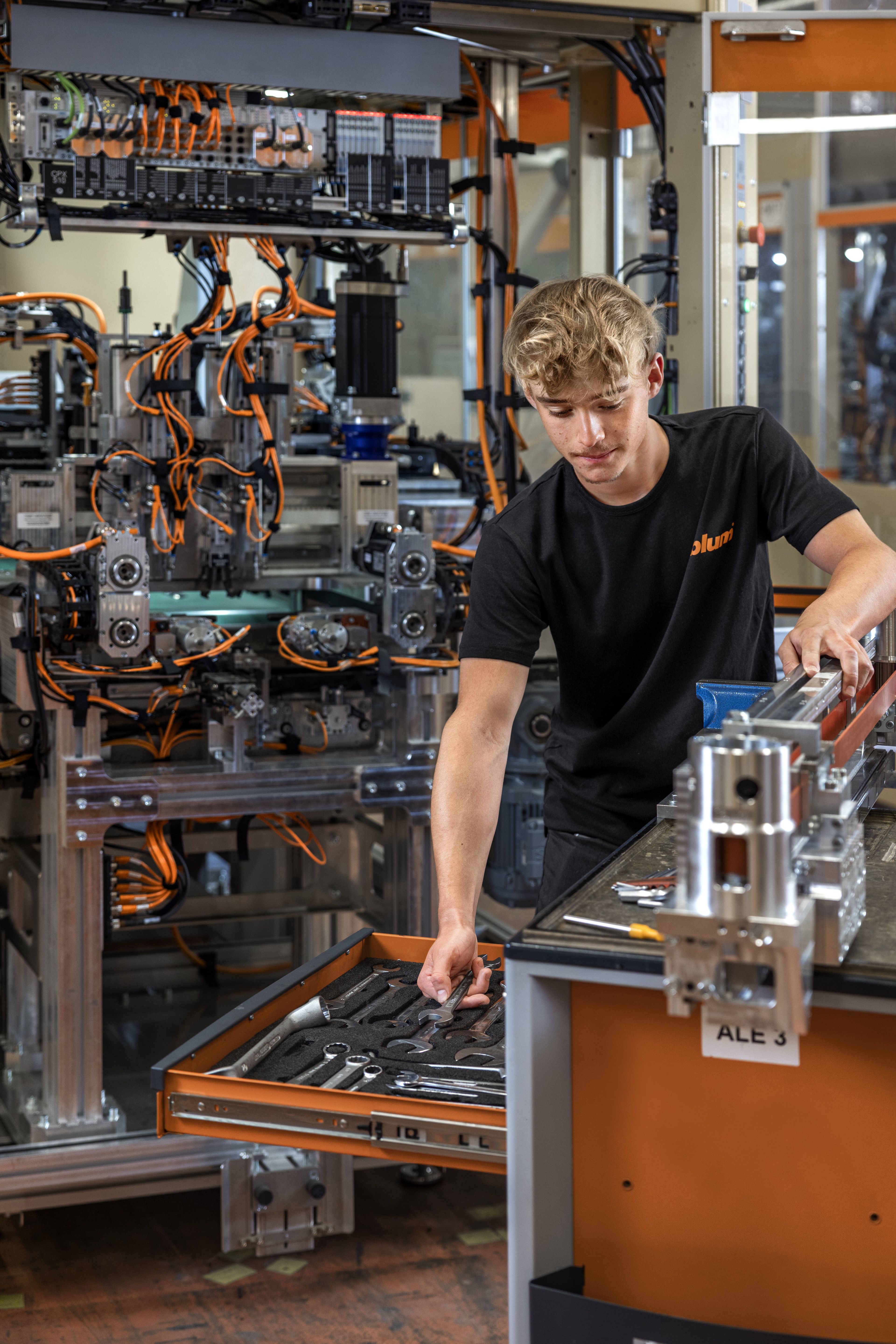 Das Bild zeigt einen Lehrling der Maschinenbautechnik, der gerade ein Förderband montiert, dafür verwendet er unterschiedliches Werkzeug aus seinem Werkzeugwagen. 