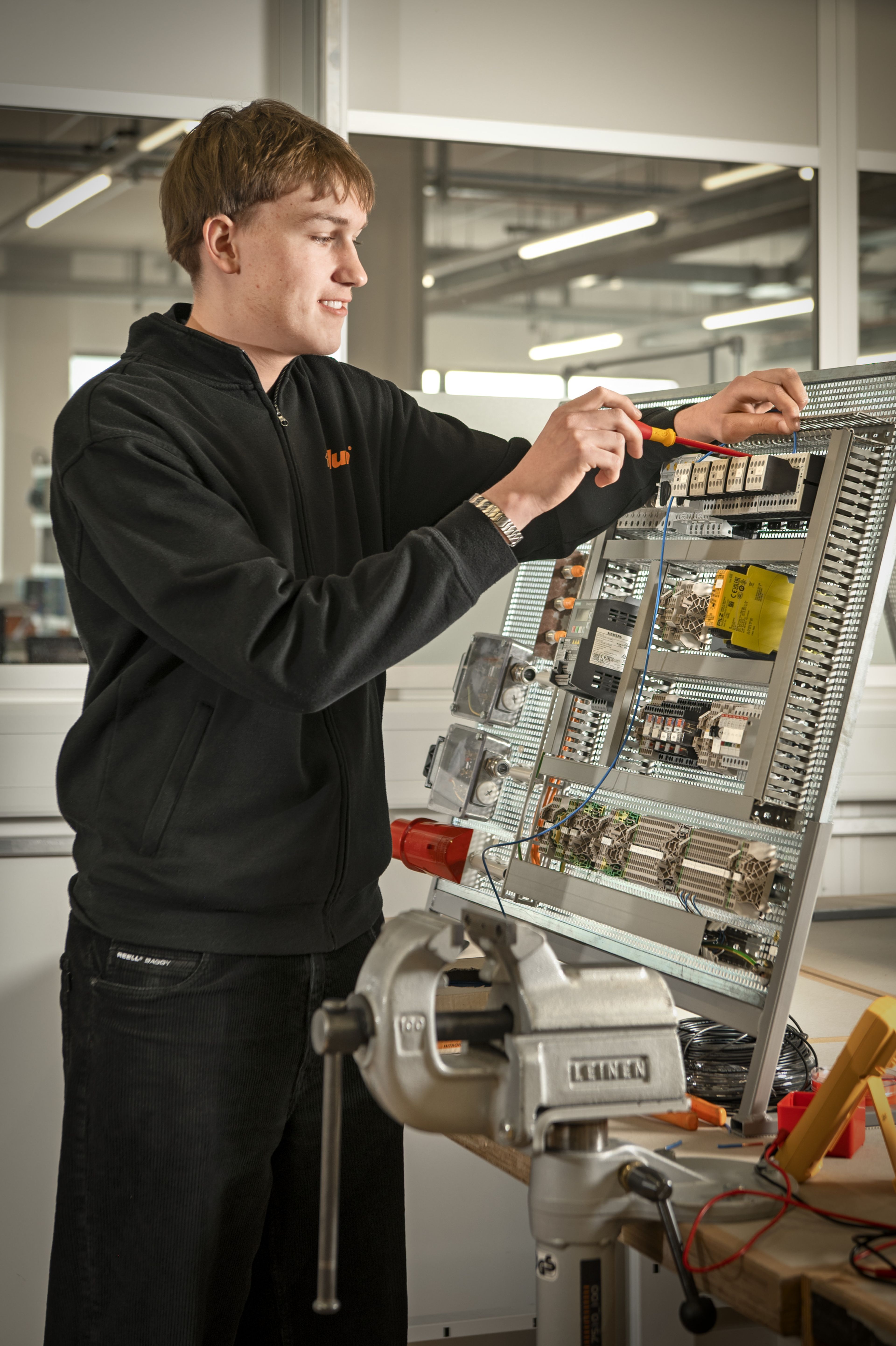 Auszubildender im Bereich Elektrotechnik bei Blum montiert und verdrahtet mit einem Schraubenzieher eine elektrische Steuerung an einem aufgebauten Schaltschrank in einer modernen Ausbildungswerkstatt.