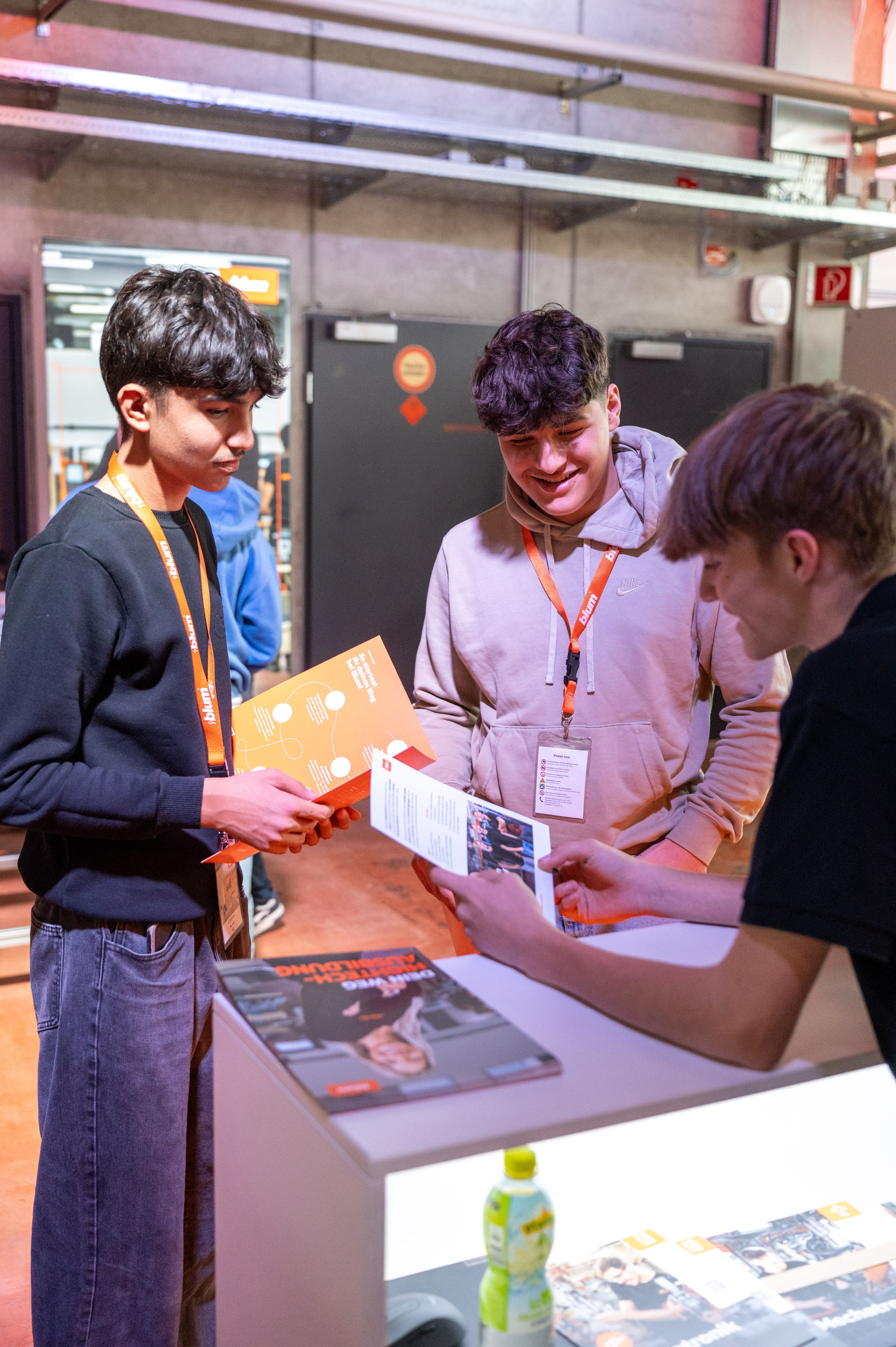 Jugendliche informieren sich an einem Messestand auf der Ausbildungsmesse über Ausbildungsberufe bei Blum. Auf dem Stand liegen Informationsbroschüren und Flyer zur Lehre, während eine Person Inhalte erläutert. Die Ausbildungsmesse bietet Einblicke in technische und kaufmännische Berufsausbildungen sowie Karrierechancen für Jugendliche.