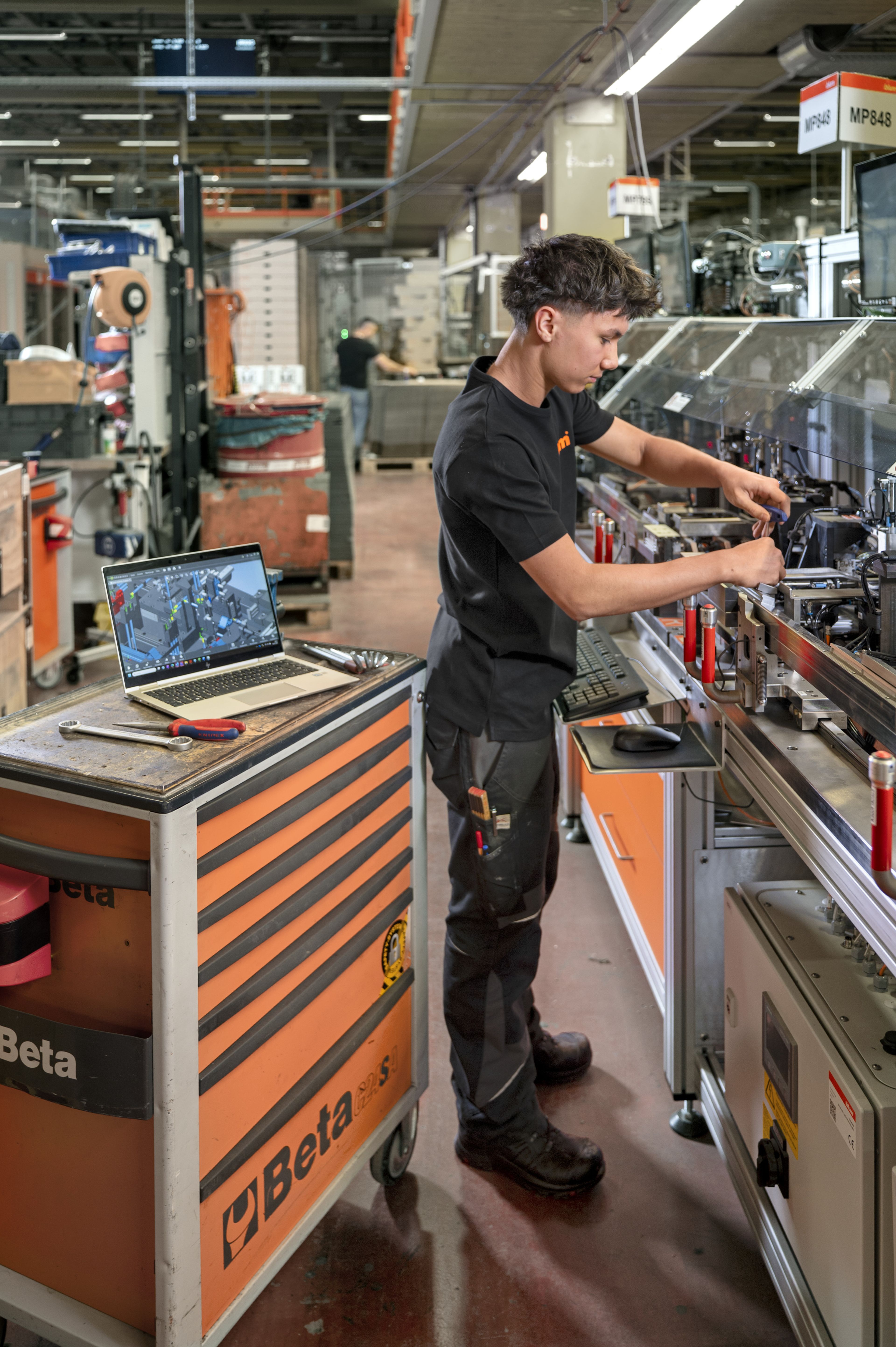 Das Bild zeigt einen Lehrling der Maschinenbautechnik, der gerade ein Förderband montiert, dafür verwendet er unterschiedliches Werkzeug aus seinem Werkzeugwagen. 