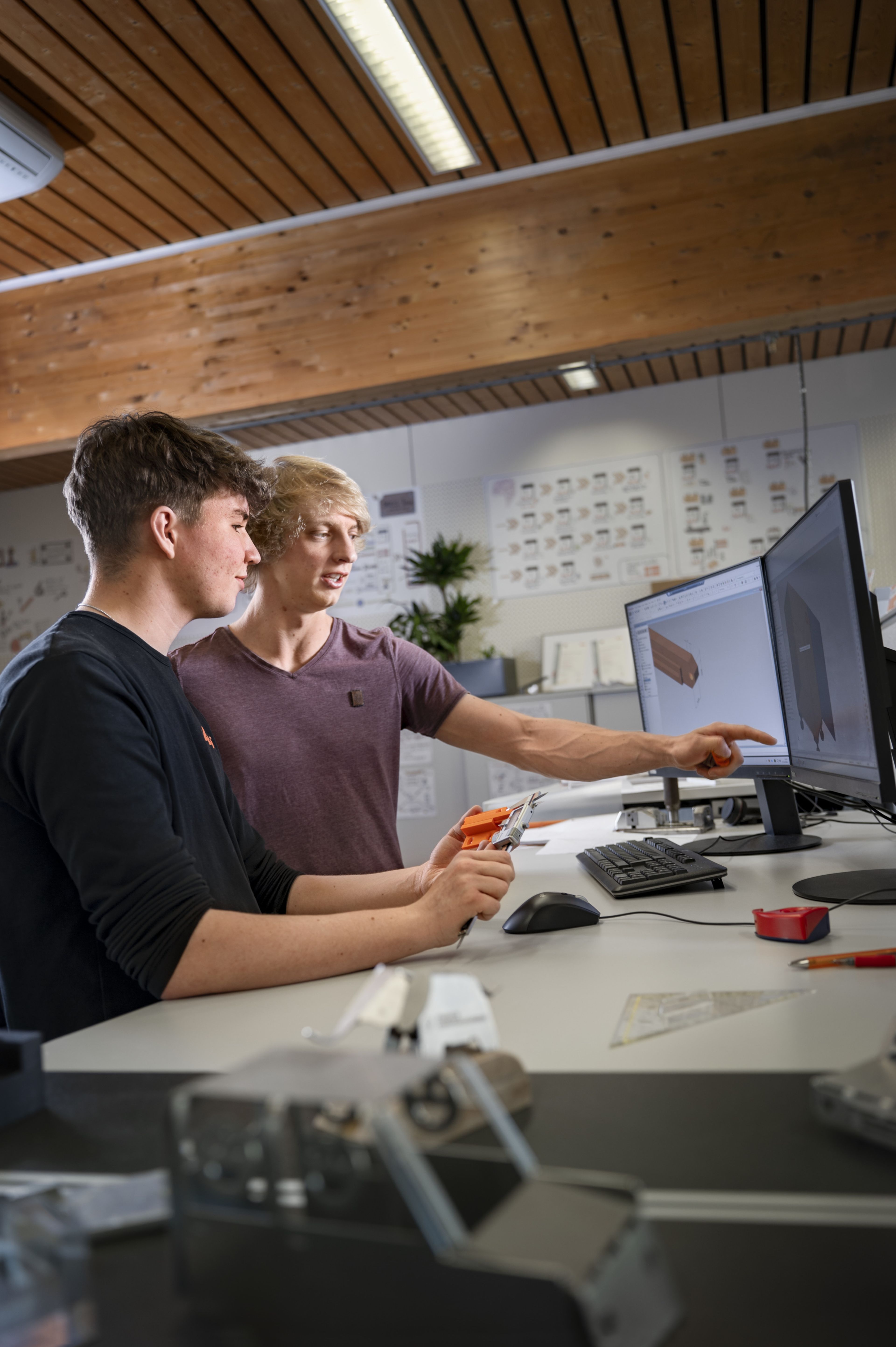 Zwei Personen arbeiten gemeinsam an einem CAD‑Entwurf in der Konstruktionstechnik und betrachten 3D‑Modelle von Bauteilen auf zwei Monitoren. Die Szene zeigt die Planung von Werkzeugen und Maschinen im technischen Arbeitsumfeld.