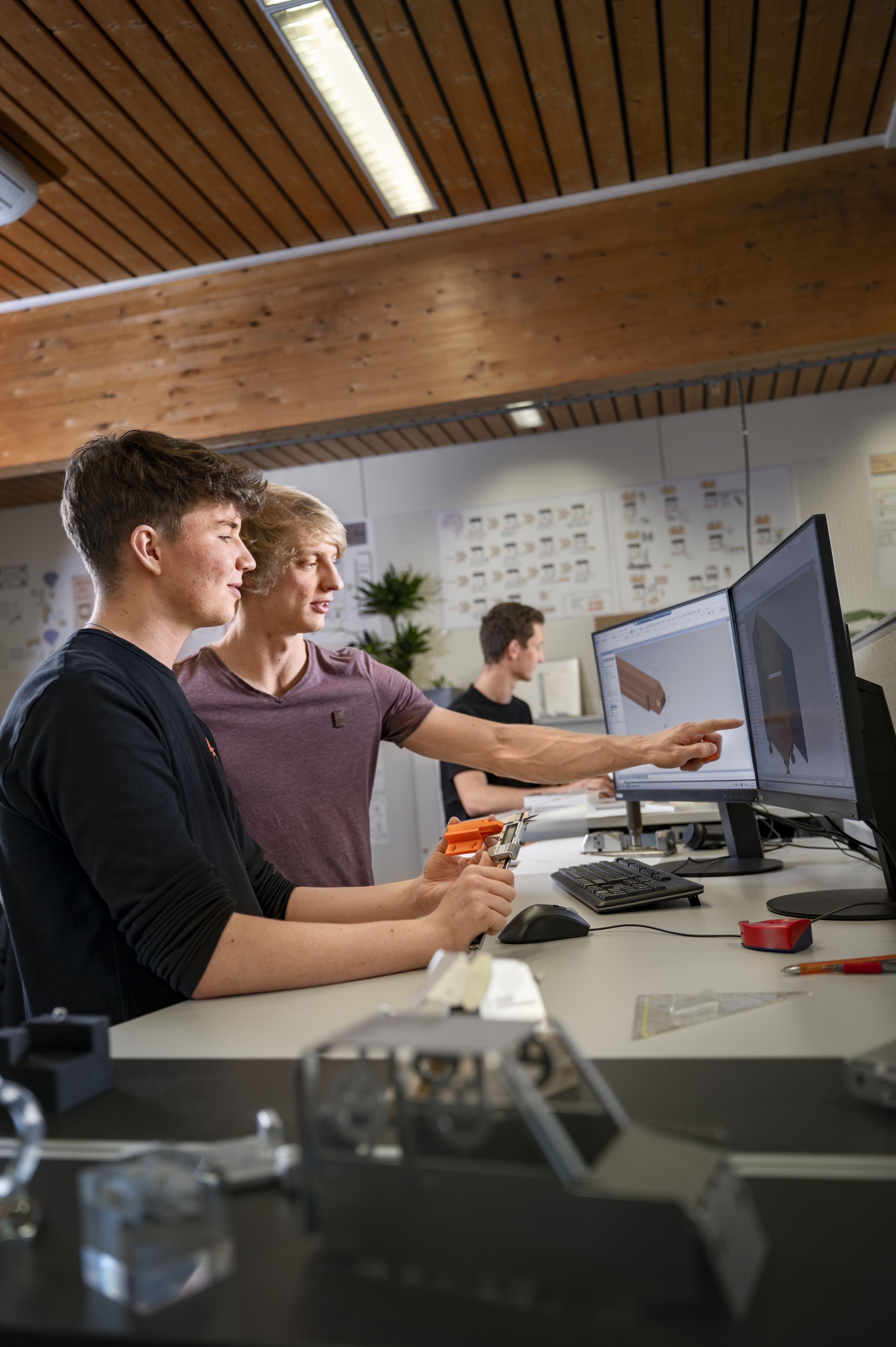 Studierende der Dualen Akademie Konstruktionstechnik analysieren ein CAD‑Modell an einem Computerarbeitsplatz bei Blum. Konstruktionsteile werden händisch vermessen und als 3D‑gedruckte Modelle visualisiert, um technische Lösungen aus Werkzeug‑ und Maschinenbau praxisnah zu überprüfen.
