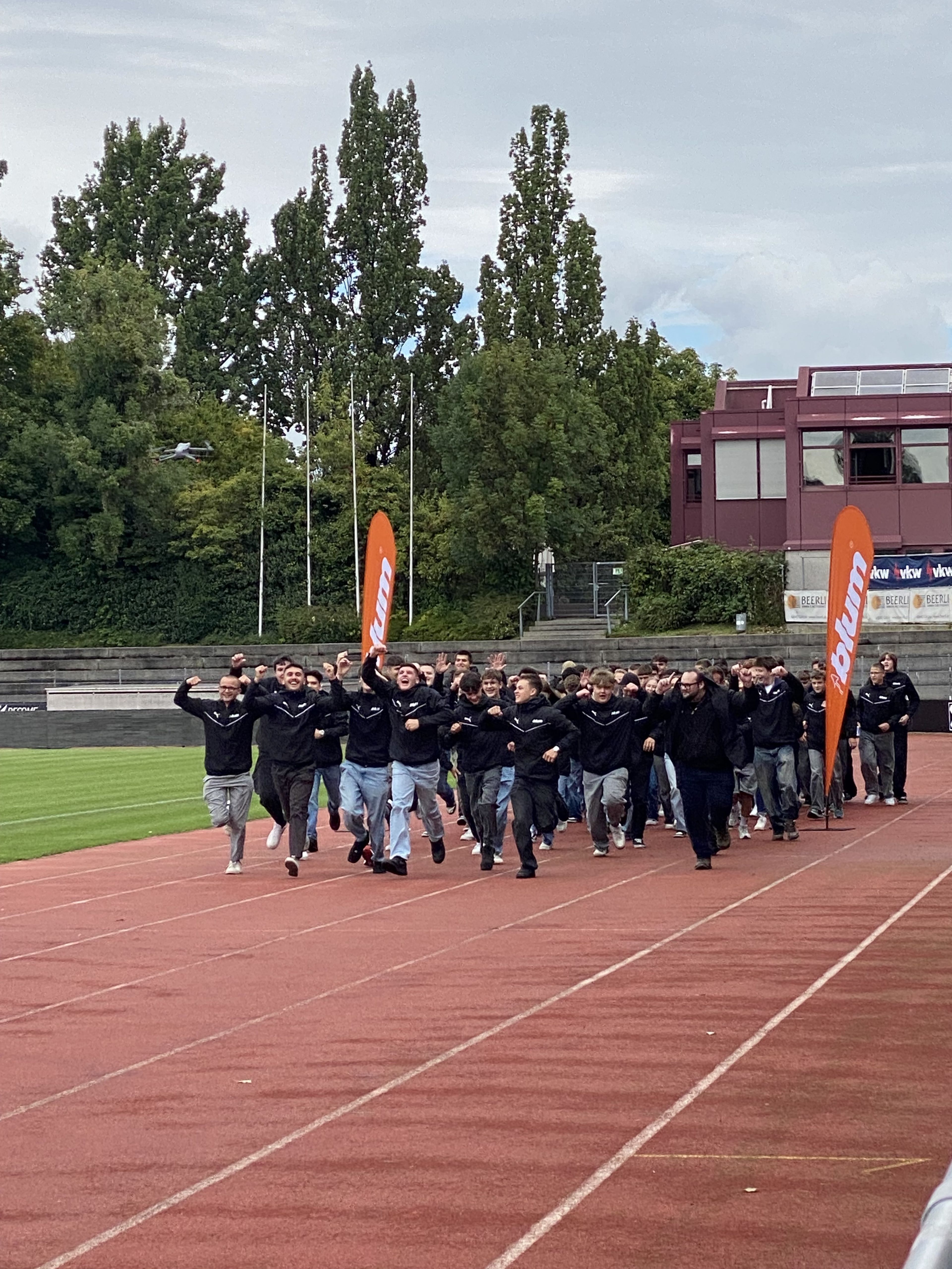 Eine Gruppe von jungen, neuen Auszubildenden steht in einer Einfahrt. Alle tragen schwarze Blum-Jacken und eine orange Blum-Sonnenbrille. Sie lachen und jubeln.