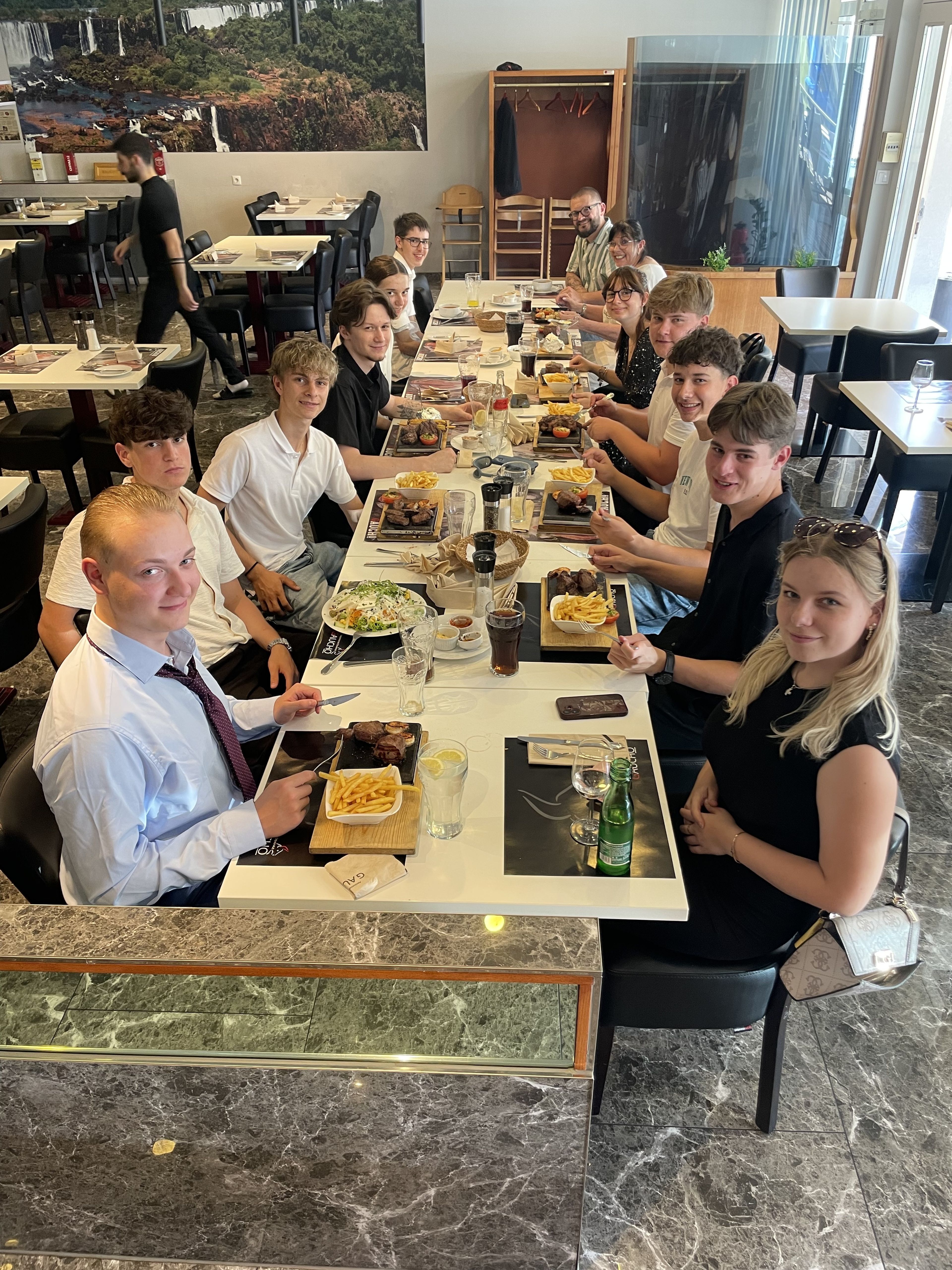 Die erste Blum-Maturaklasse feiert nach der erfolgreich bestandenen Englisch-Matura 2025 gemeinsam in einem Steakhouse. Elf Schülerinnen und Schüler sitzen an einer gedeckten Tafel und genießen das gemeinsame Abschlussessen.