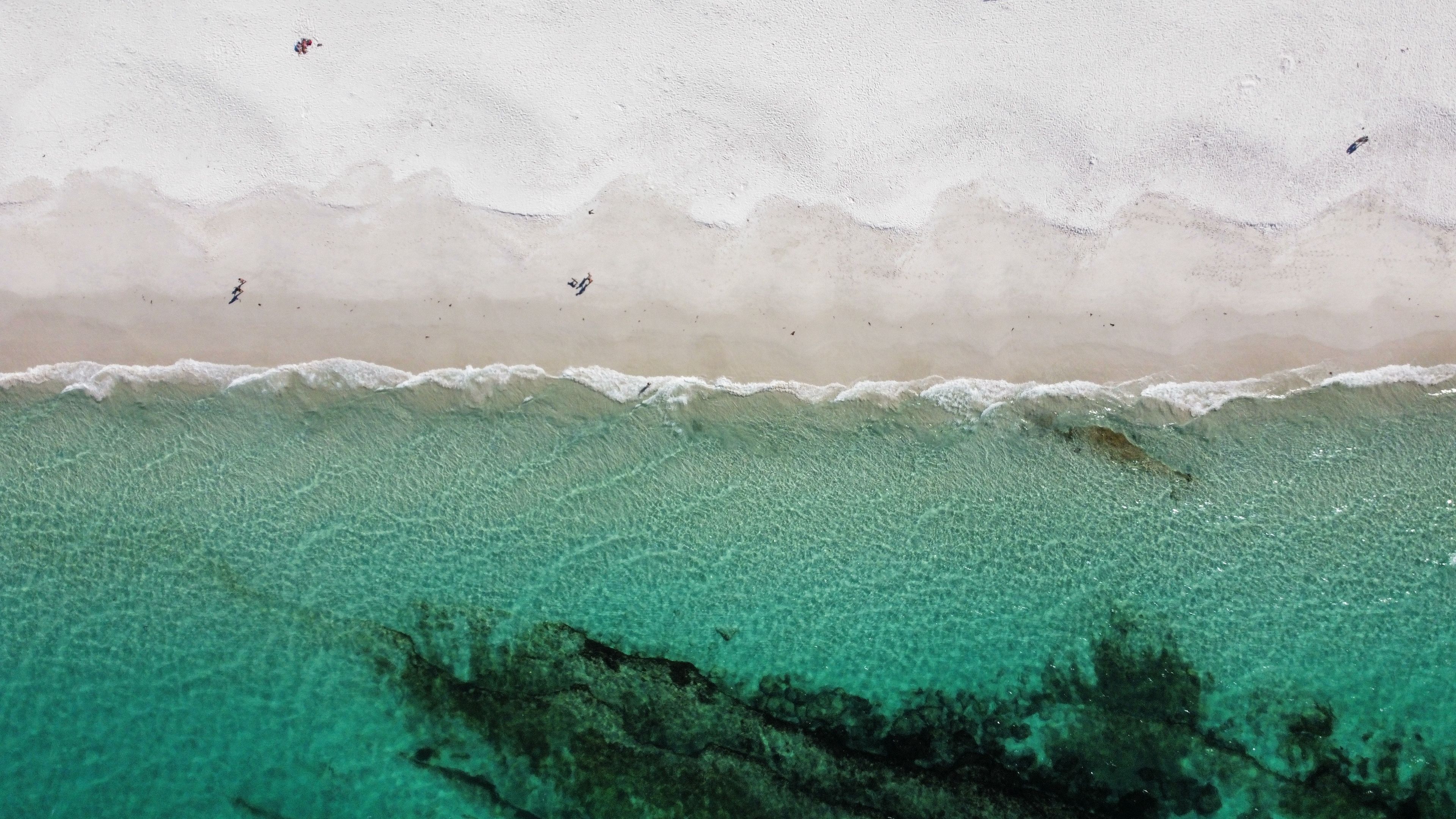 Luftaufnahme eines Strandes mit weißem Sand, sanft plätschernden Wellen und klarem, türkisfarbenem Wasser