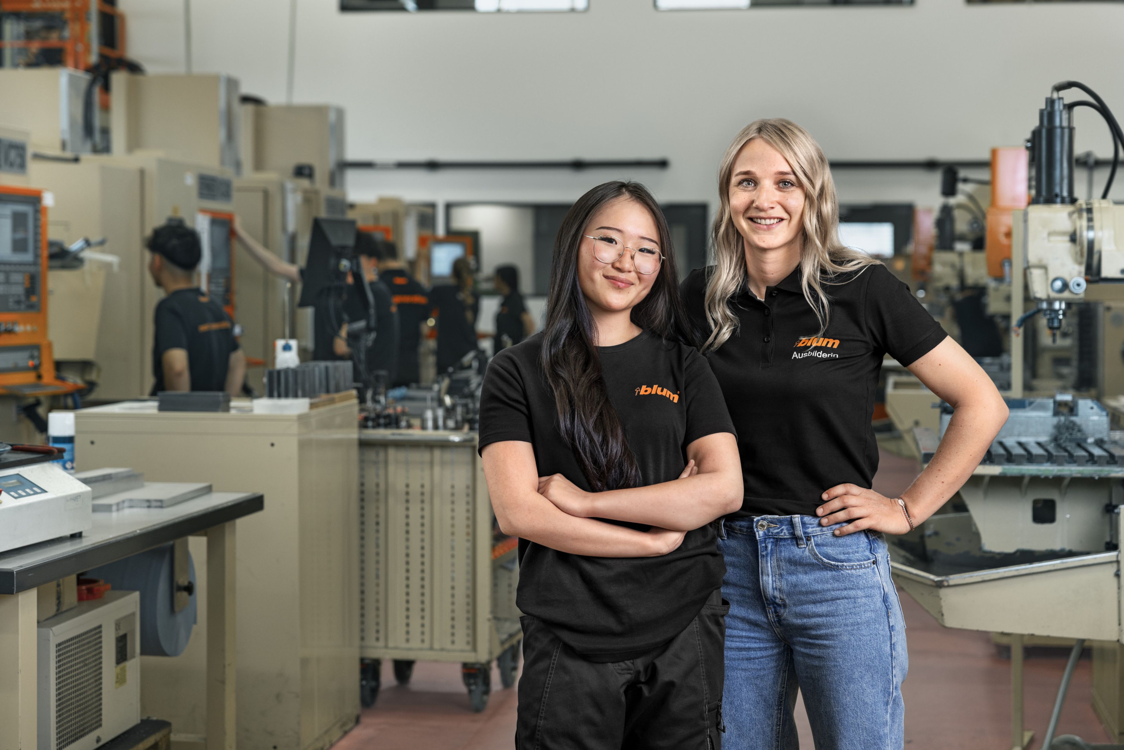 Zwei Frauen in schwarzer Arbeitskleidung mit Blum‑Logo stehen selbstbewusst in einer Produktionsumgebung der Metall‑Grundausbildung bei Blum. Das Bild zeigt Frauen in der Ausbildung anlässlich des Weltfrauentags am 8. März; aktuell absolvieren 87 junge Frauen ihre Ausbildung bei Blum.