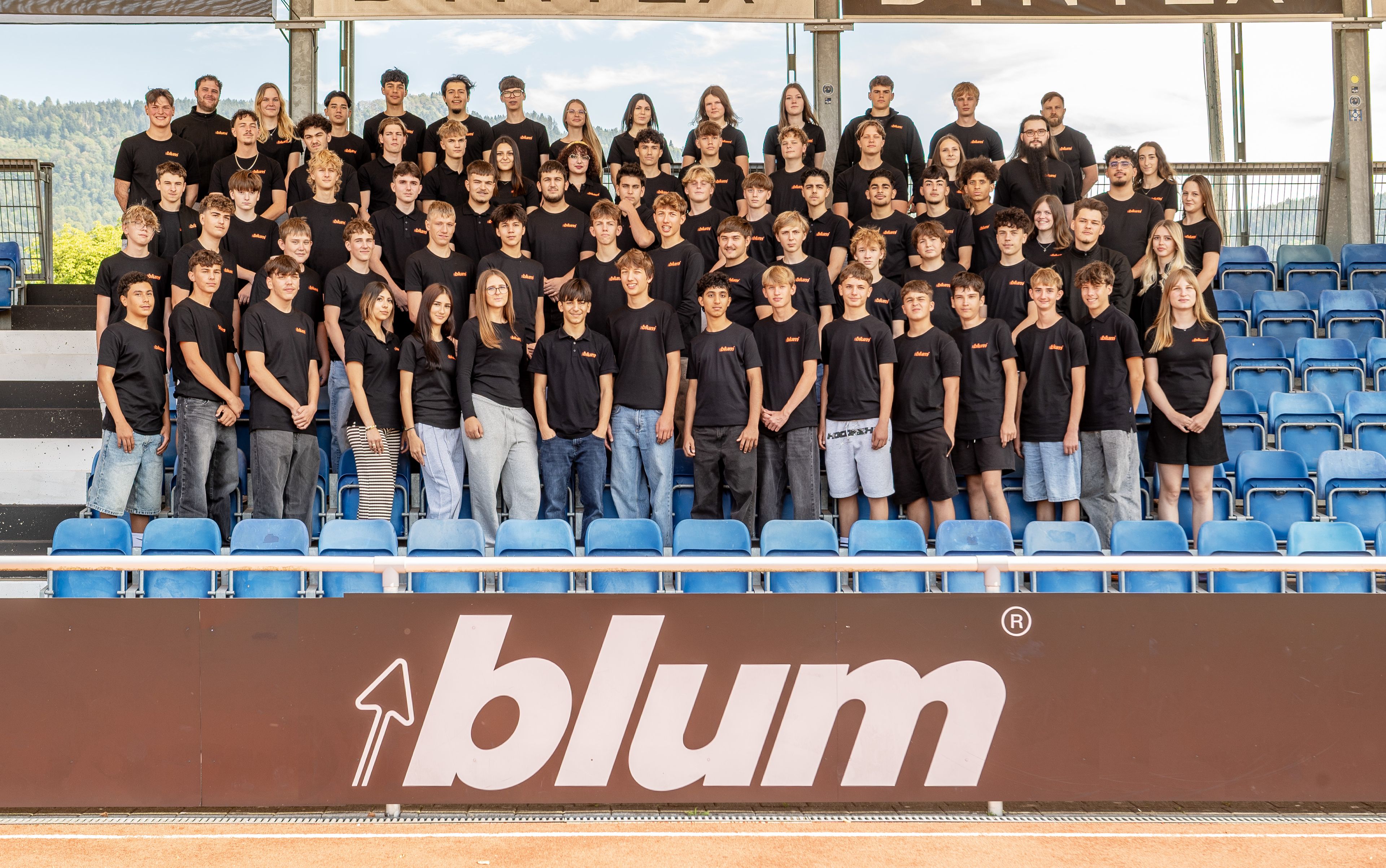 Gruppenfoto der 76 neuen Auszubildenden von Blum in Vorarlberg zum Ausbildungsstart am 1. September. Die Lehrlinge stehen gemeinsam auf einer Tribüne vor einem Blum-Schriftzug.