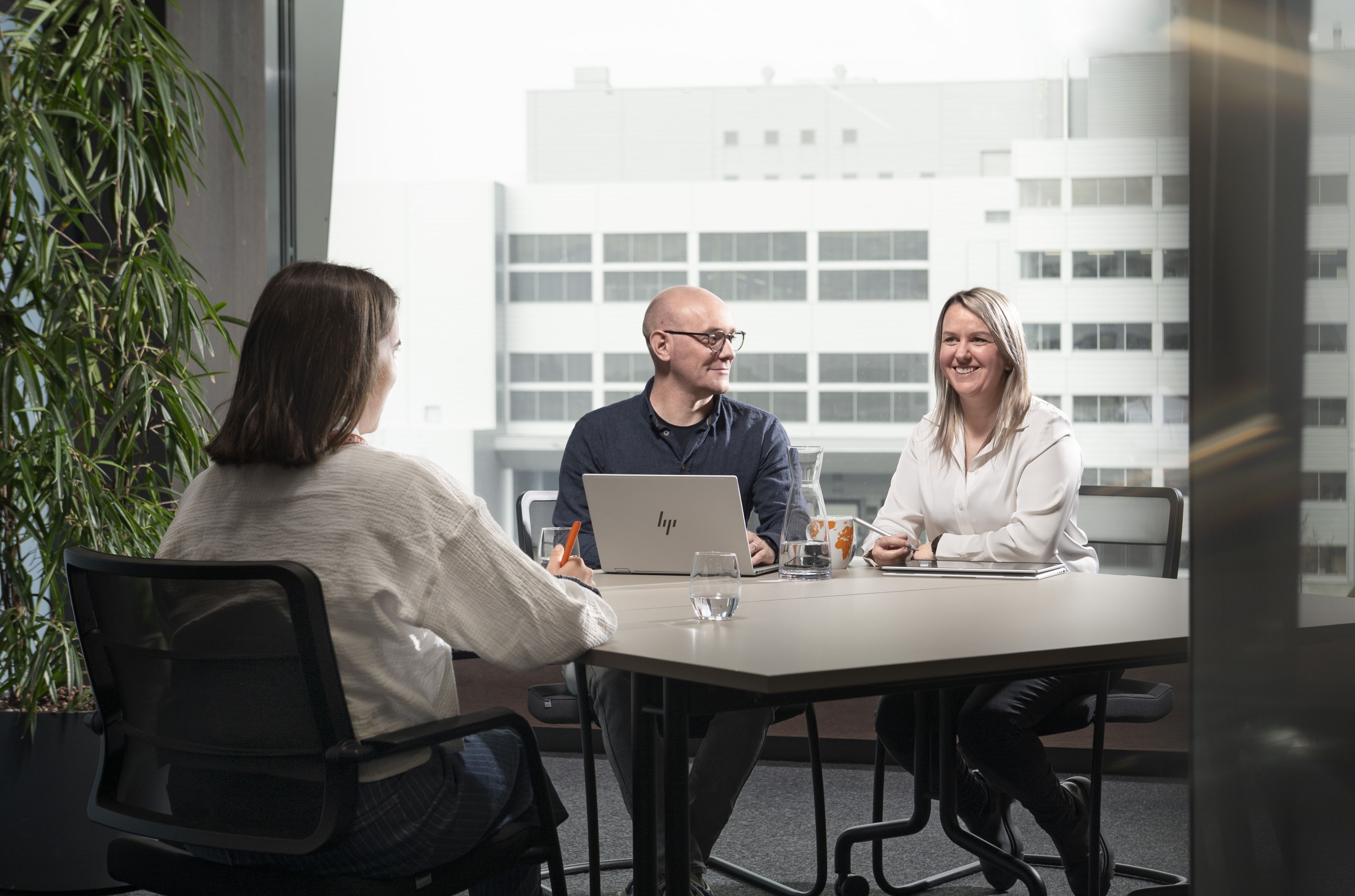 Three people at a meeting table in an office, with a laptop open, engaged in conversation, and a large window view of a building outside.