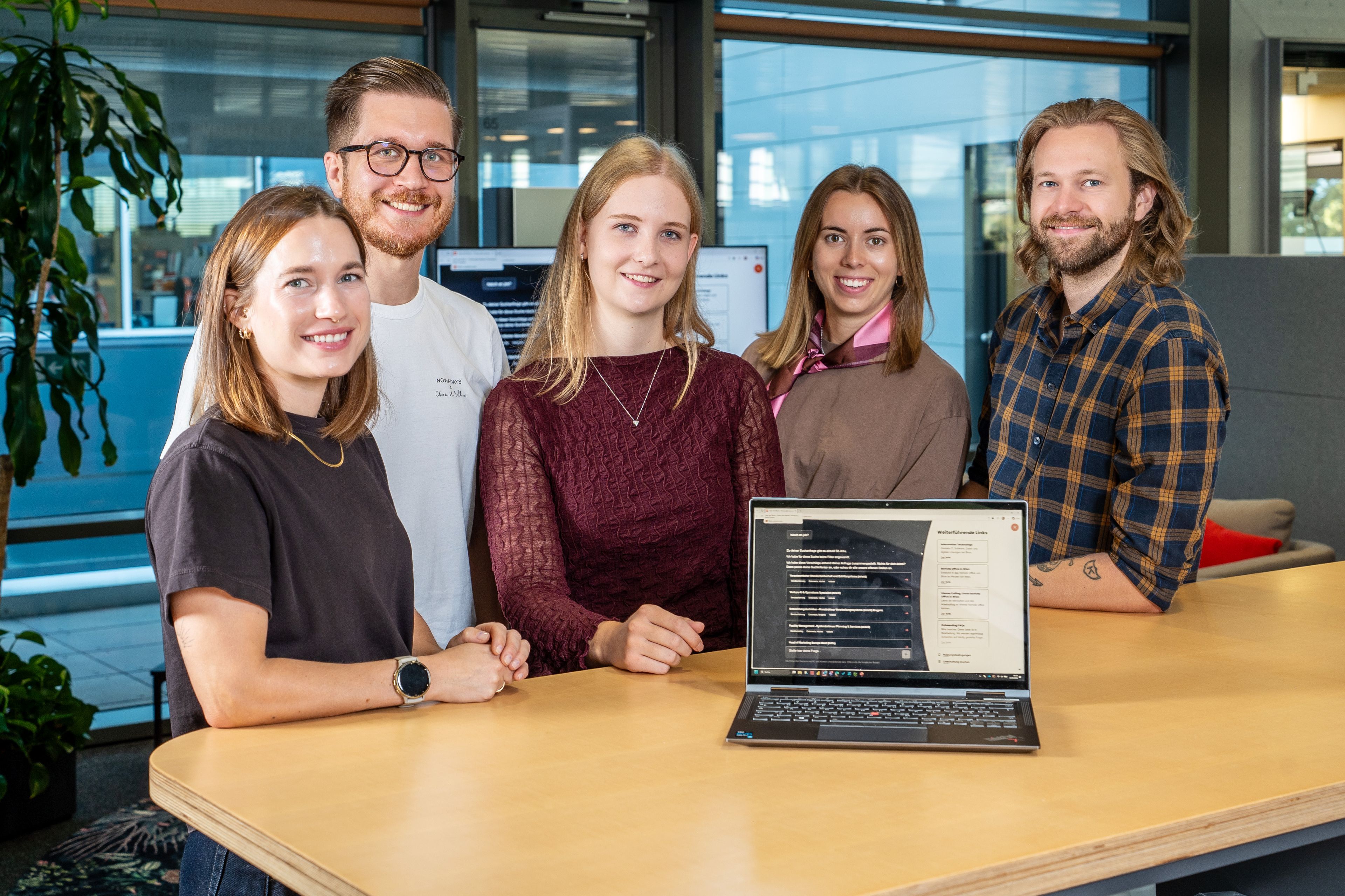 Five people stand around a table in a modern office, with a laptop on the table displaying a website.