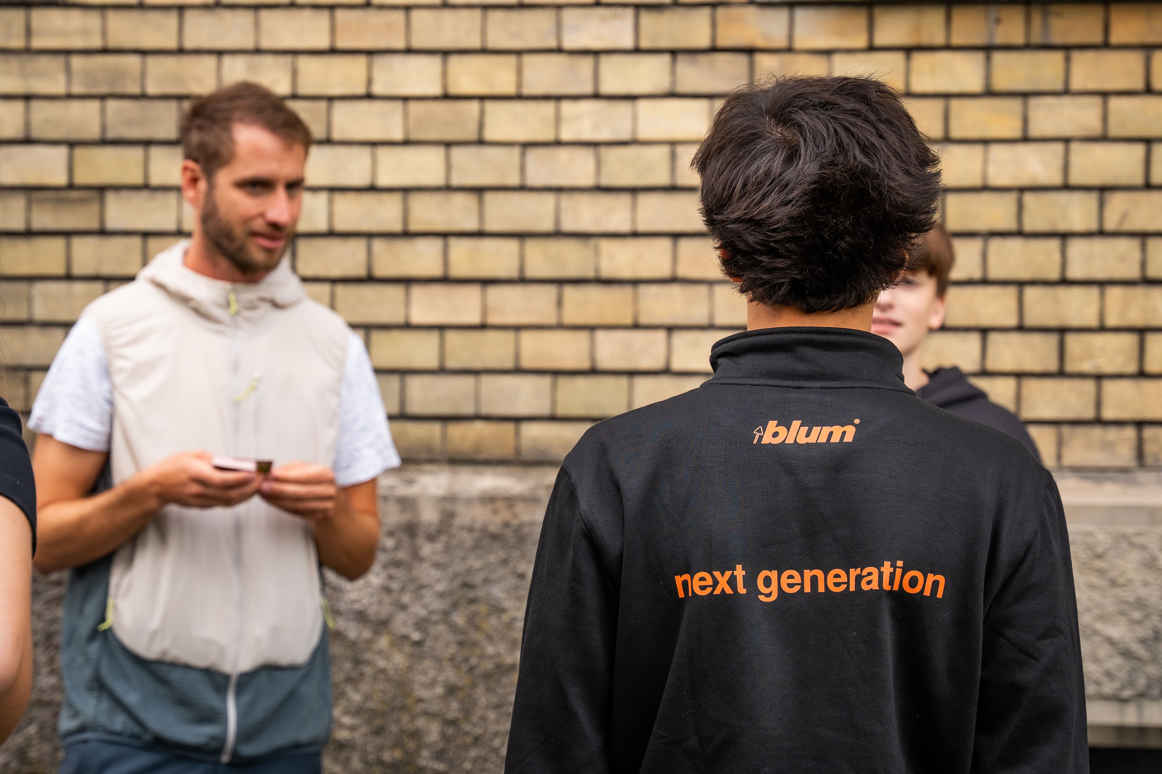 Ausbildungsstart bei Blum: Neue Auszubildende nehmen am Startworkshop teil. Im Vordergrund ist eine Person mit einer Jacke mit der Aufschrift „next generation“ zu sehen, während weitere Teilnehmende vor einer Backsteinwand miteinander ins Gespräch kommen.