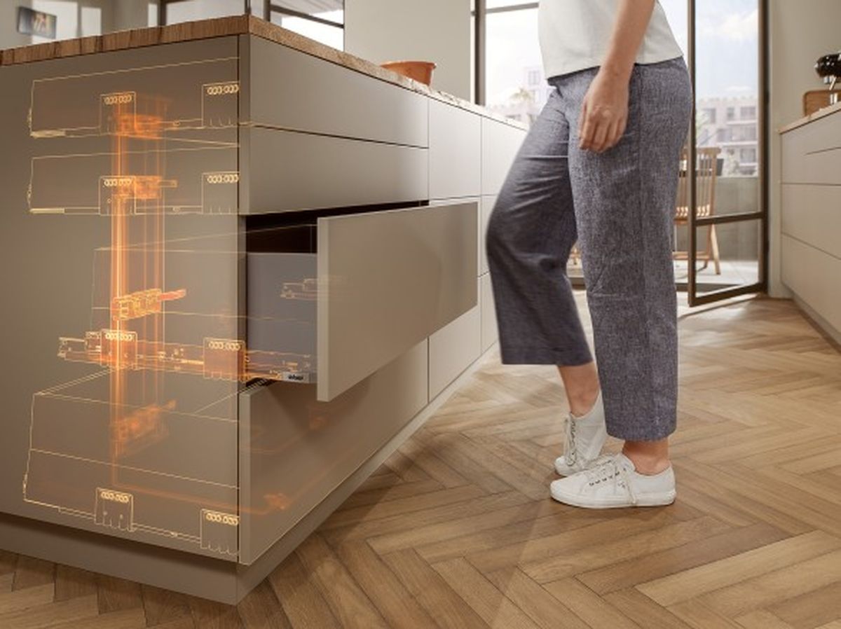 Eine Frau schließt mit dem Knie eine Blum-Schublade. Im Möbel sind in Orange die technischen Finessen der Führungen und Boxsysteme hervorgehoben.