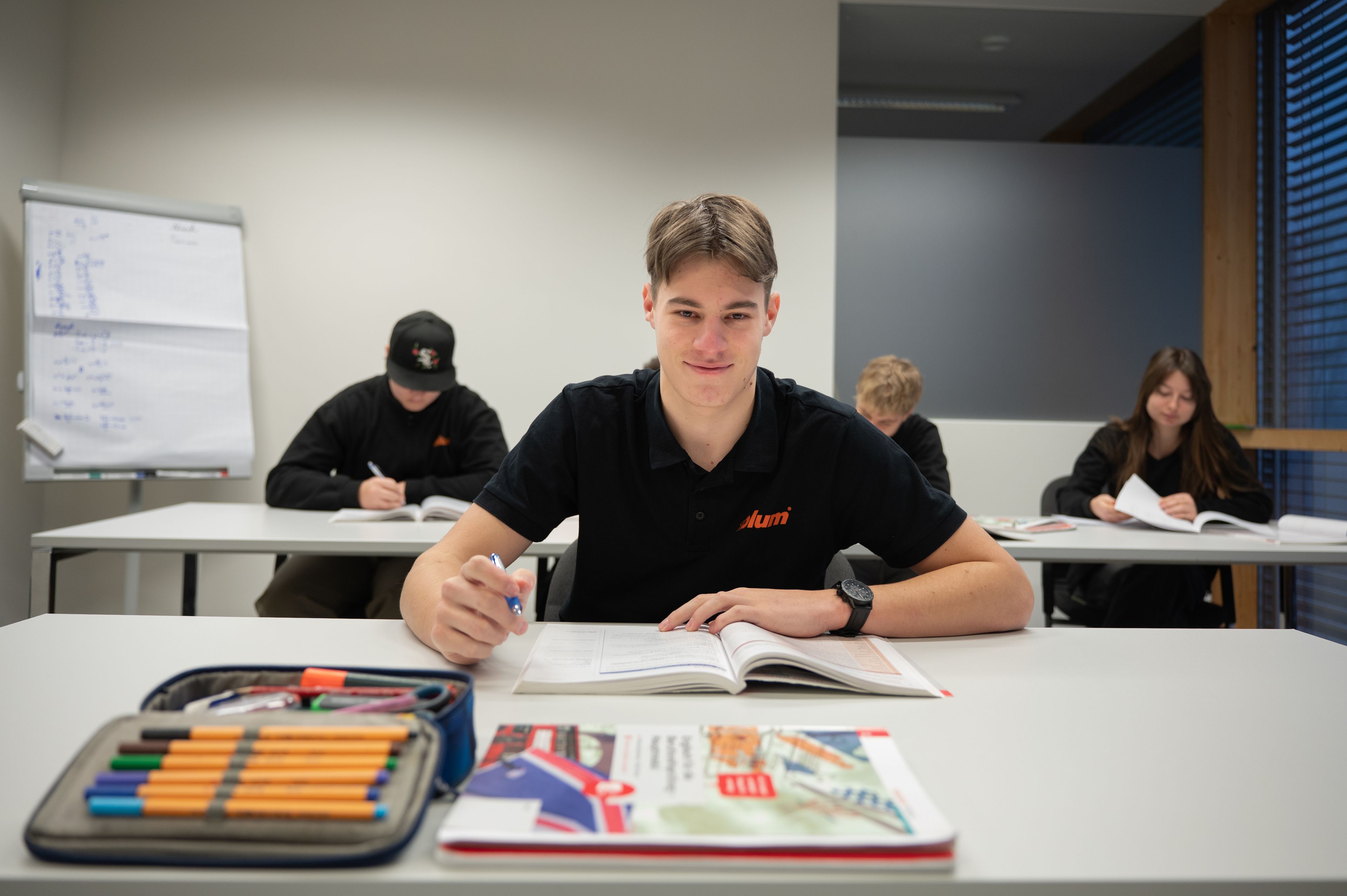 Teilnehmer der Hightech-Ausbildung mit Matura im Fachbereich CAD-Konstruktion sitzt im Schulungsraum an einem Tisch und arbeitet mit Lehrmaterialien. Im Vordergrund liegen bunte Stifte und ein aufgeschlagenes Buch, im Hintergrund weitere Auszubildende bei der Arbeit.