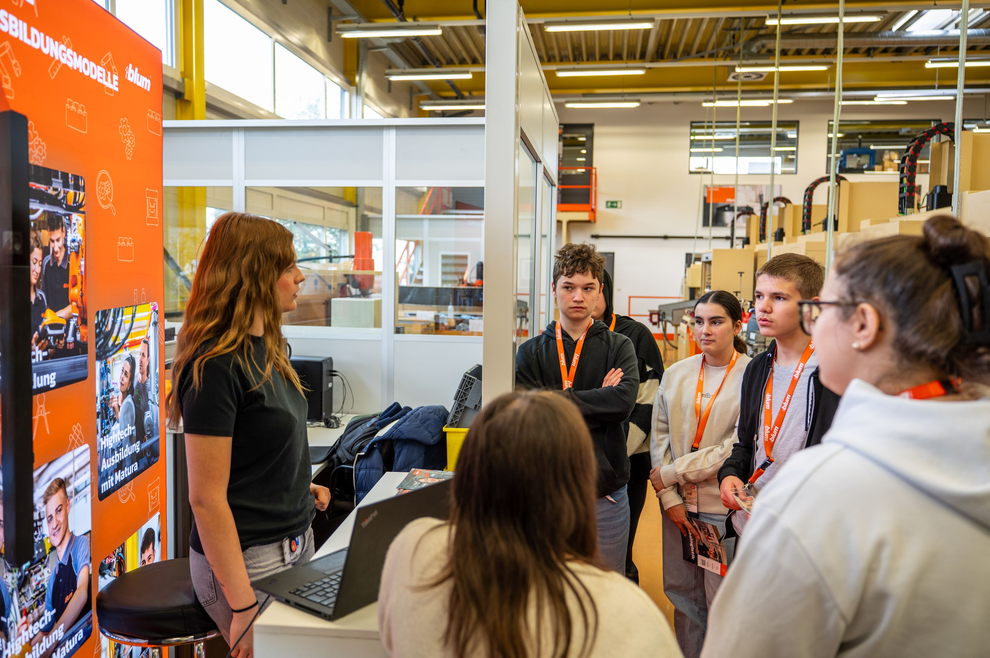 Informationsstand zur Hightech-Ausbildung bei Blum in einer Werkhalle. Eine Ausbilderin erklärt einer Gruppe von Jugendlichen die Ausbildungsmodelle, darunter Lehrberufe und die Möglichkeit einer Ausbildung mit Matura. Im Hintergrund sind Maschinen und Werkbänke sichtbar, auf dem Display stehen Infos zu Ausbildungswegen.