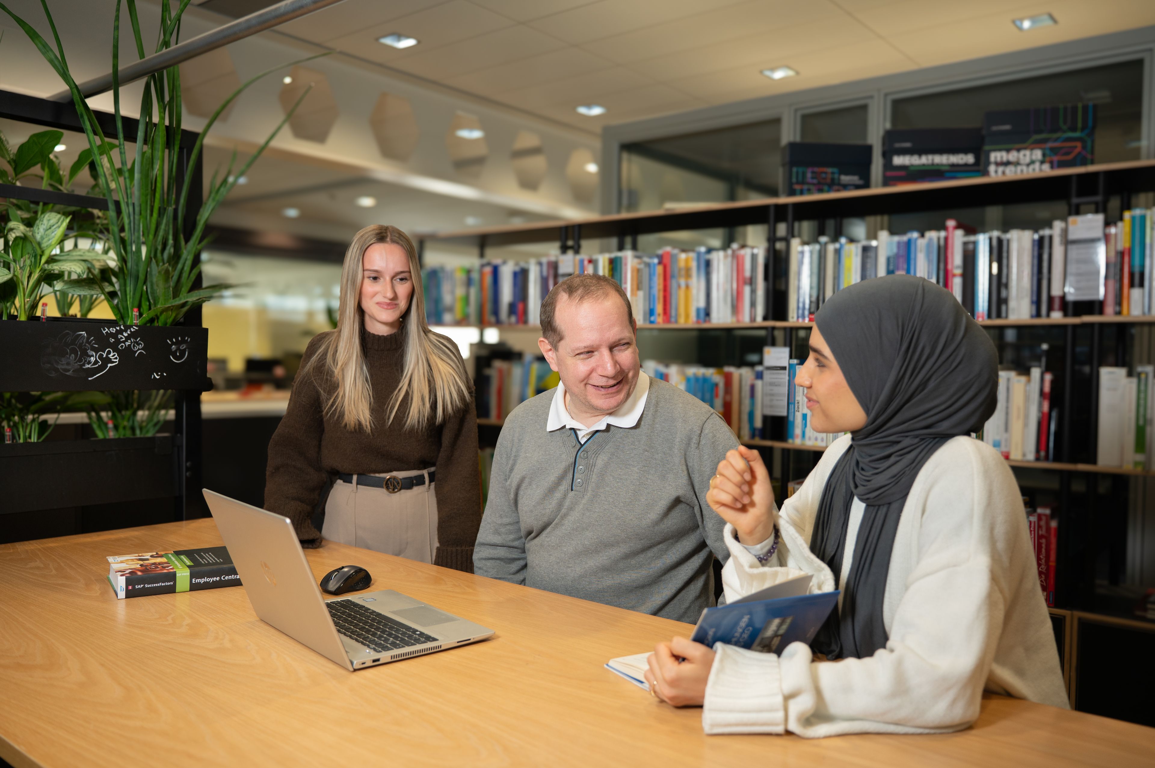 Drei Personen unterhalten sich an einem Tisch in einer Bibliothek mit einem Laptop, umgeben von Bücherregalen und Pflanzen.