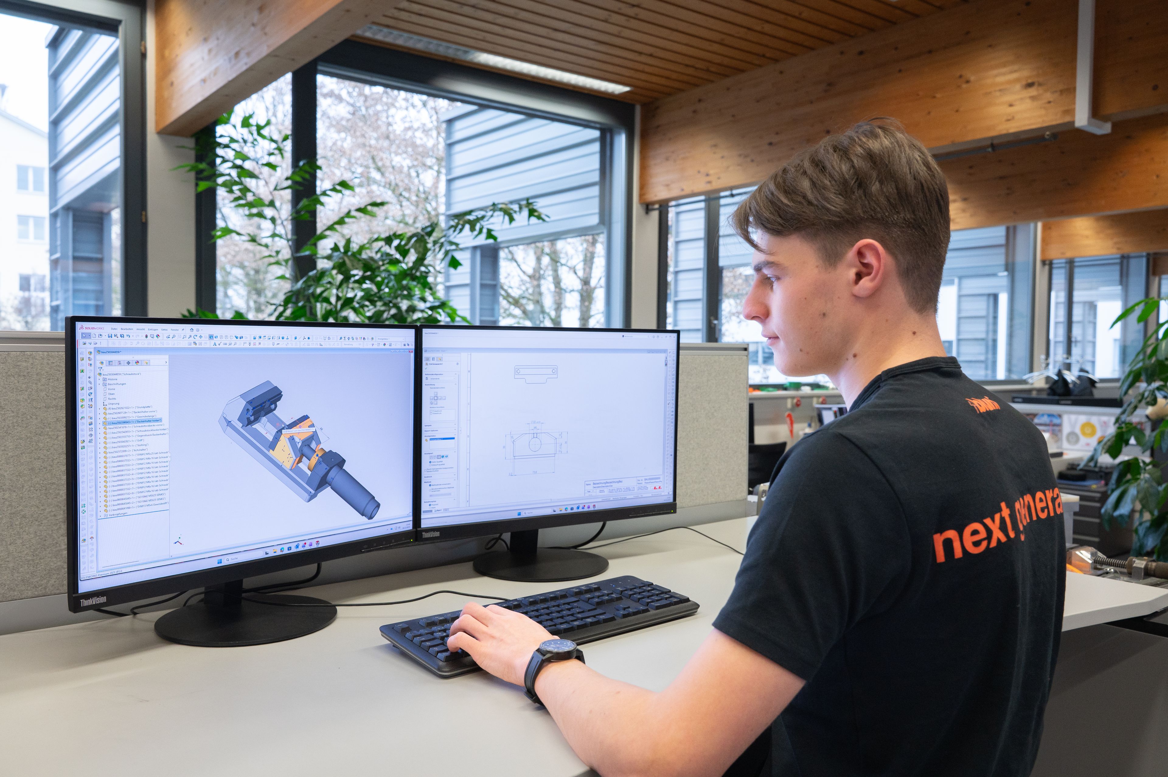 Auszubildender der Hightech-Ausbildung mit Matura im Fachbereich CAD-Konstruktion arbeitet an einem Arbeitsplatz mit zwei Monitoren. Auf den Bildschirmen sind 3D-CAD-Modelle und technische Zeichnungen zu sehen. Im Hintergrund große Fenster mit Blick auf moderne Bürogebäude.