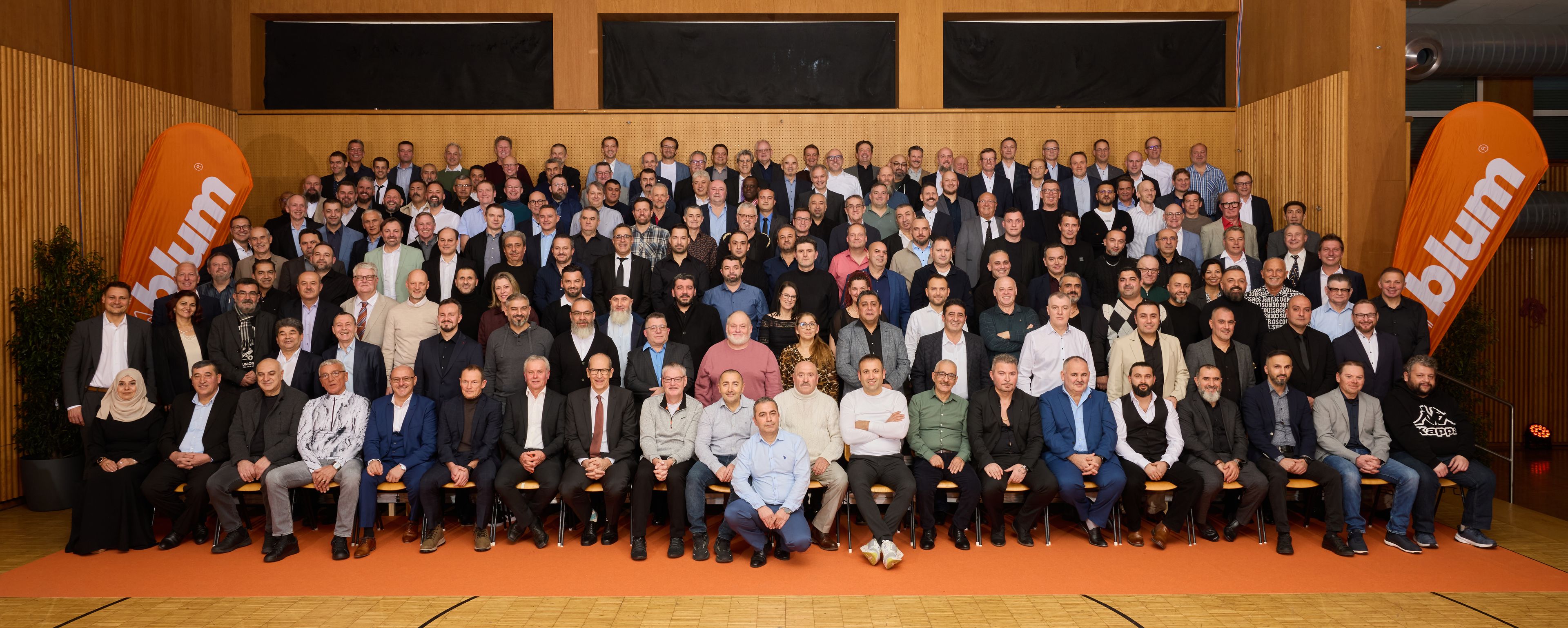 Eine große Gruppe von Menschen posiert in einer Halle, an deren Seiten zwei orangefarbene „Blum“-Banner hängen; sie tragen sowohl formelle als auch legere Kleidung.