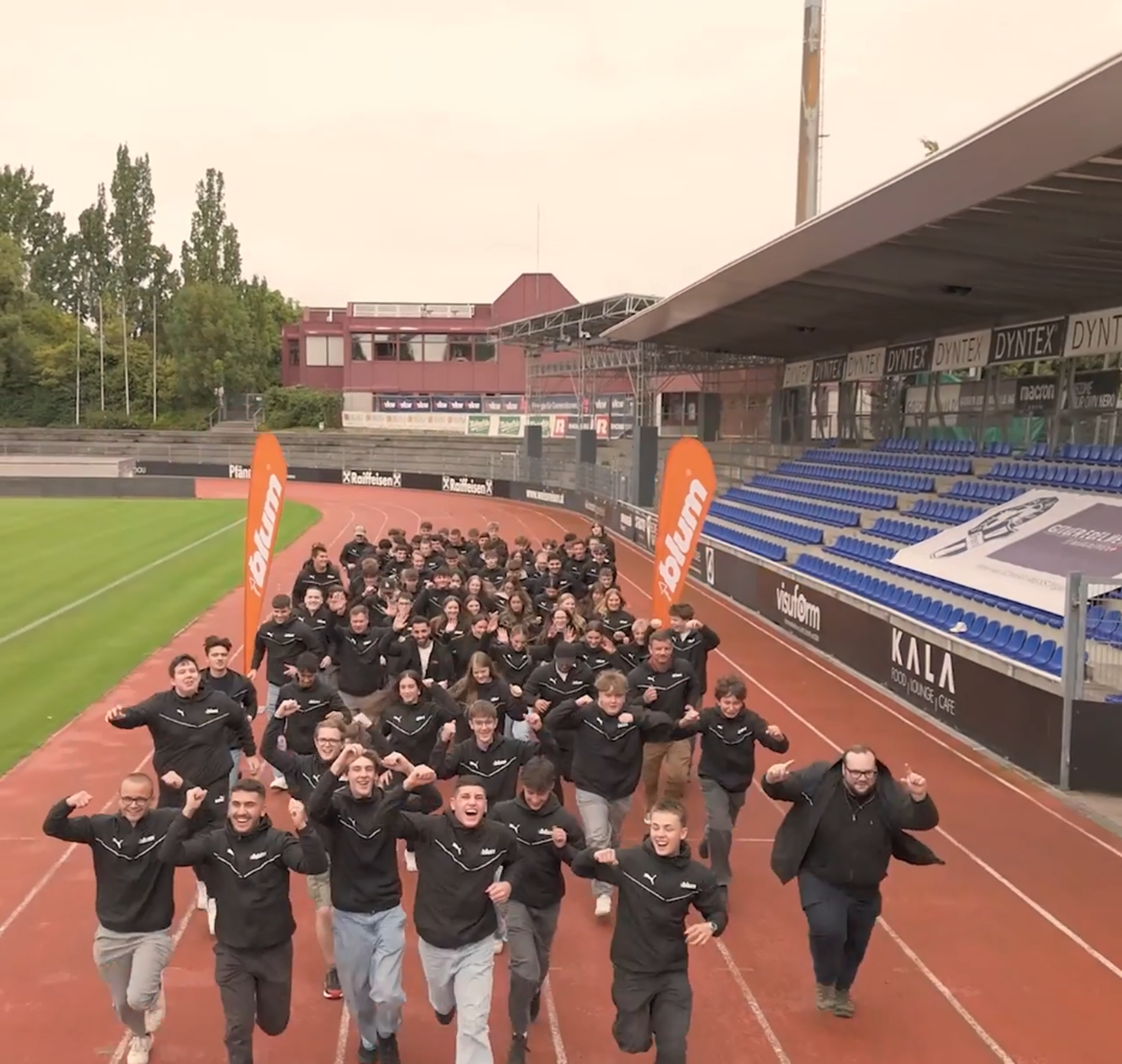 Gruppe von 71 zukünftigen Lehrlingen läuft gemeinsam über eine Stadion‑Laufbahn in Vorarlberg. Die Jugendlichen tragen schwarze Blum‑Jacken, im Hintergrund sind Tribünen und Blum‑Flaggen zu sehen. Das Bild entstand am Technikland Zusagetag Ende März, bei dem Blum Ausbildungsplätze in der Elektro‑ und Metallindustrie vergab.
