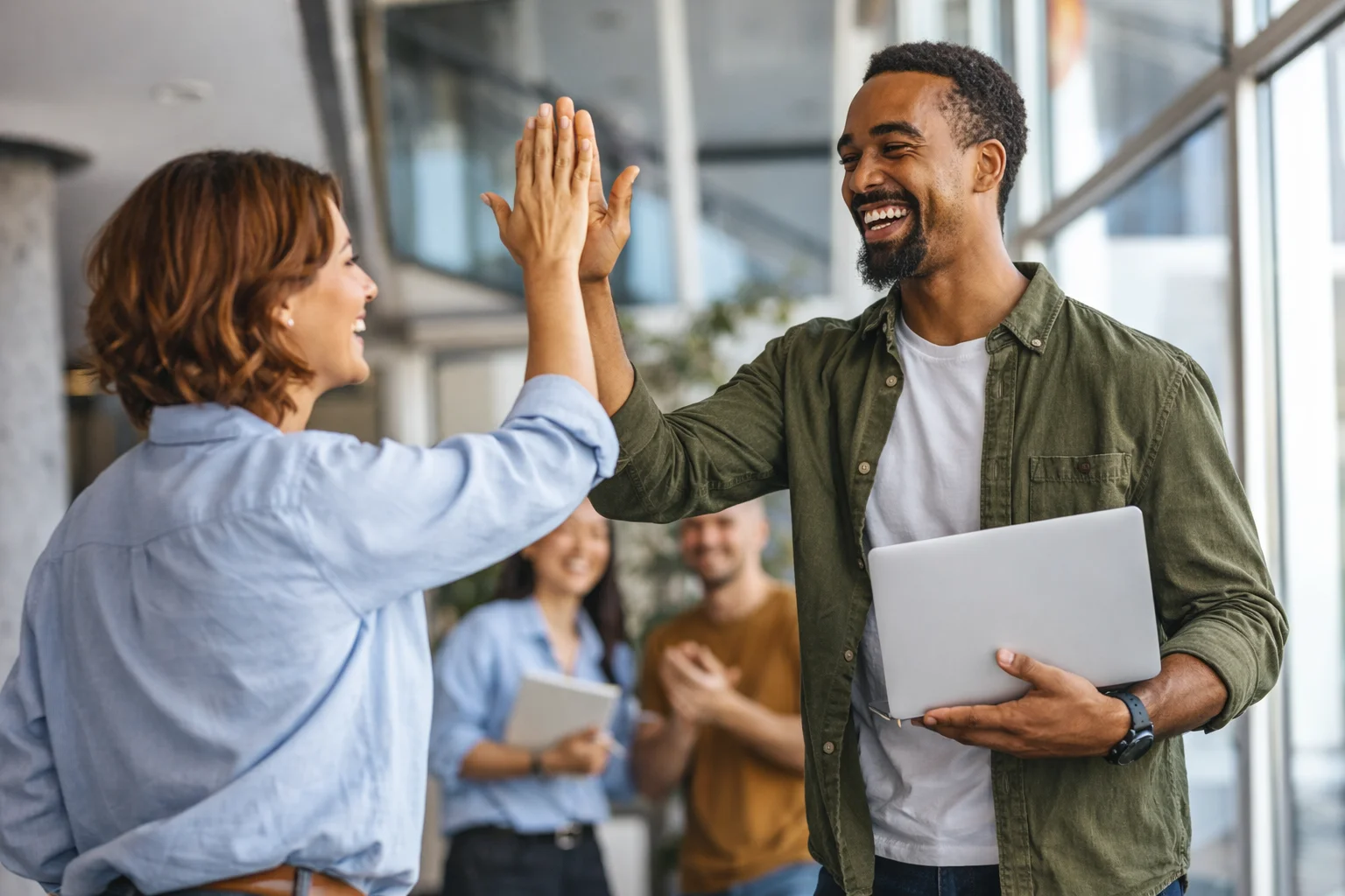 Two colleagues high-five in a modern office, smiling. One holds a laptop. Two other coworkers are in the blurred background, clapping.