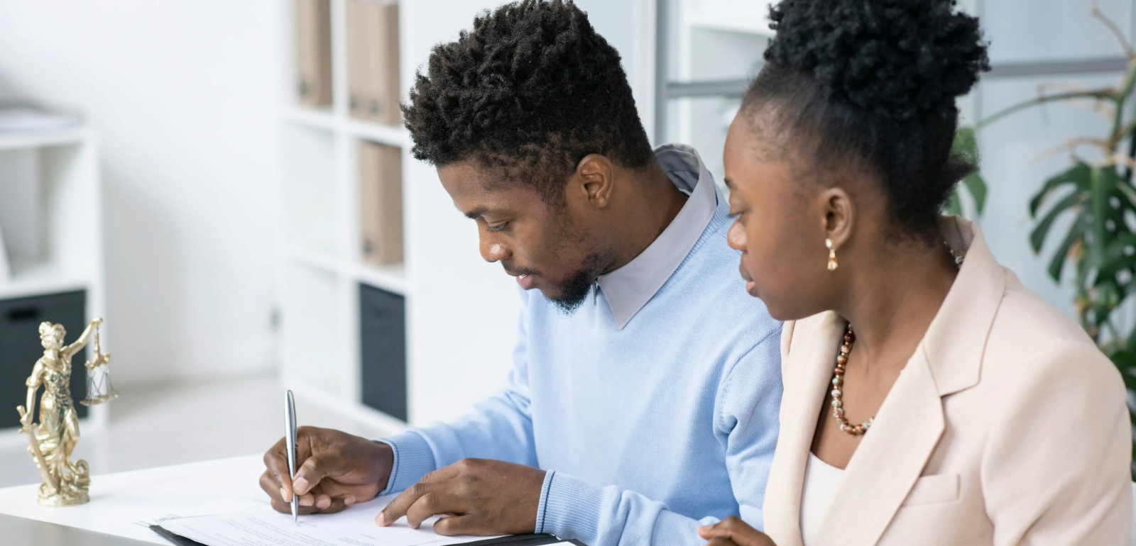 Two people seated at a desk, one writing on a document, with a small statue of Lady Justice nearby. Shelves in the background.