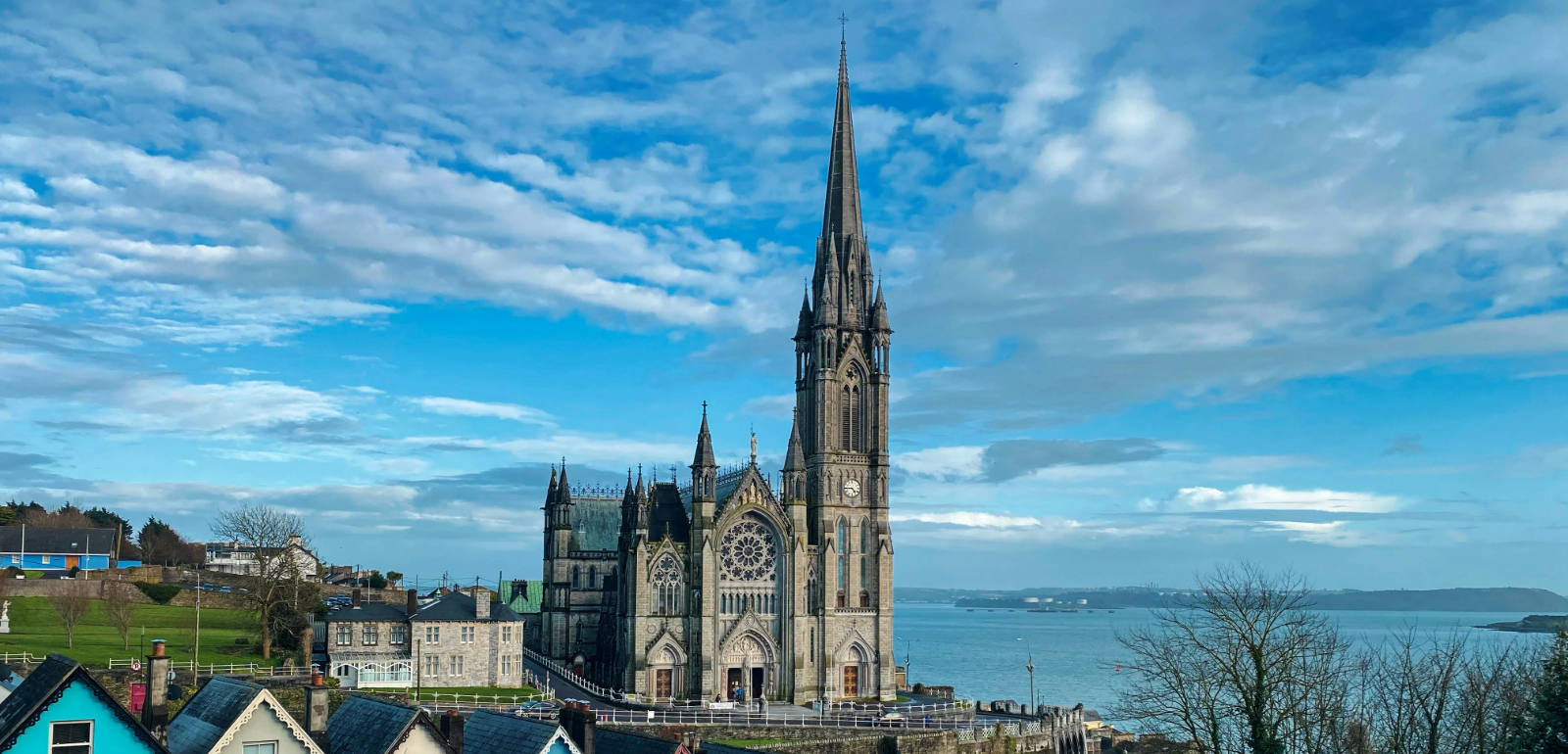 A tall, ornate Gothic cathedral with a pointed spire stands near the water, surrounded by colorful houses under a blue sky.