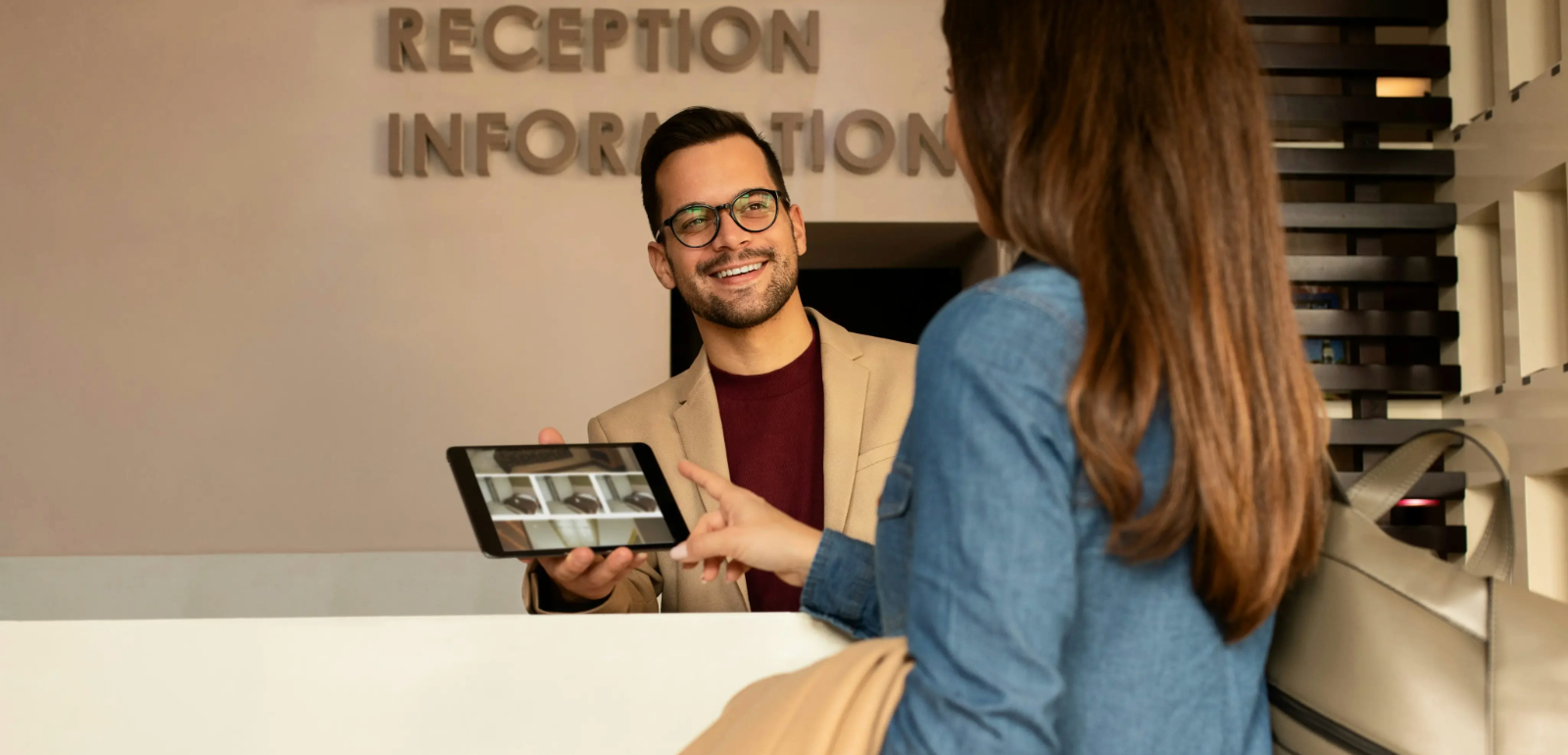 A receptionist hands a digital tablet to a woman at a counter with a sign that reads 