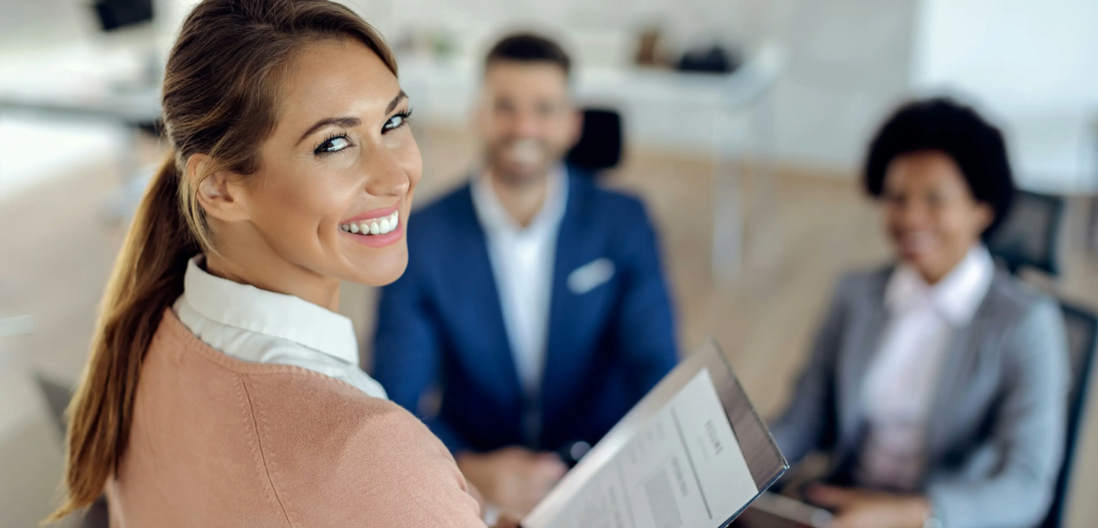 Smiling woman holding a clipboard, seated with two colleagues in a well-lit office setting.