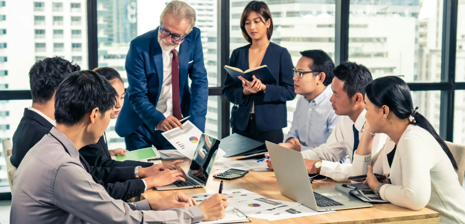 A diverse group of professionals engage in a meeting around a conference table, with charts and laptops, in a modern office setting.