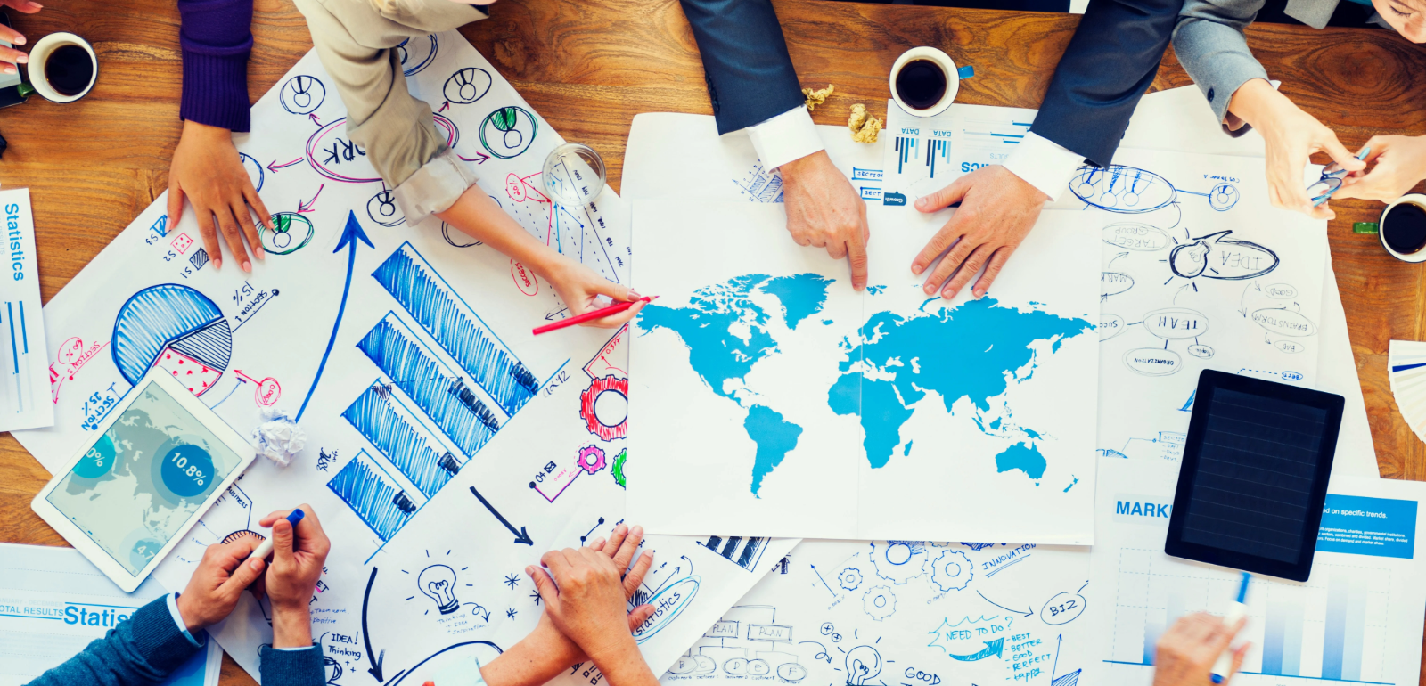 A group of people collaborates over a wooden table filled with world maps, charts, graphs, and papers, discussing strategies with coffee cups nearby.