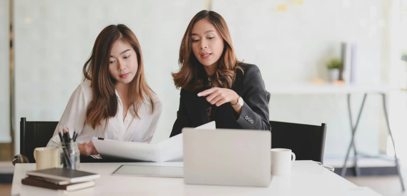Two women in an office setting, reviewing documents and discussing something, with a laptop and coffee cups on the table.