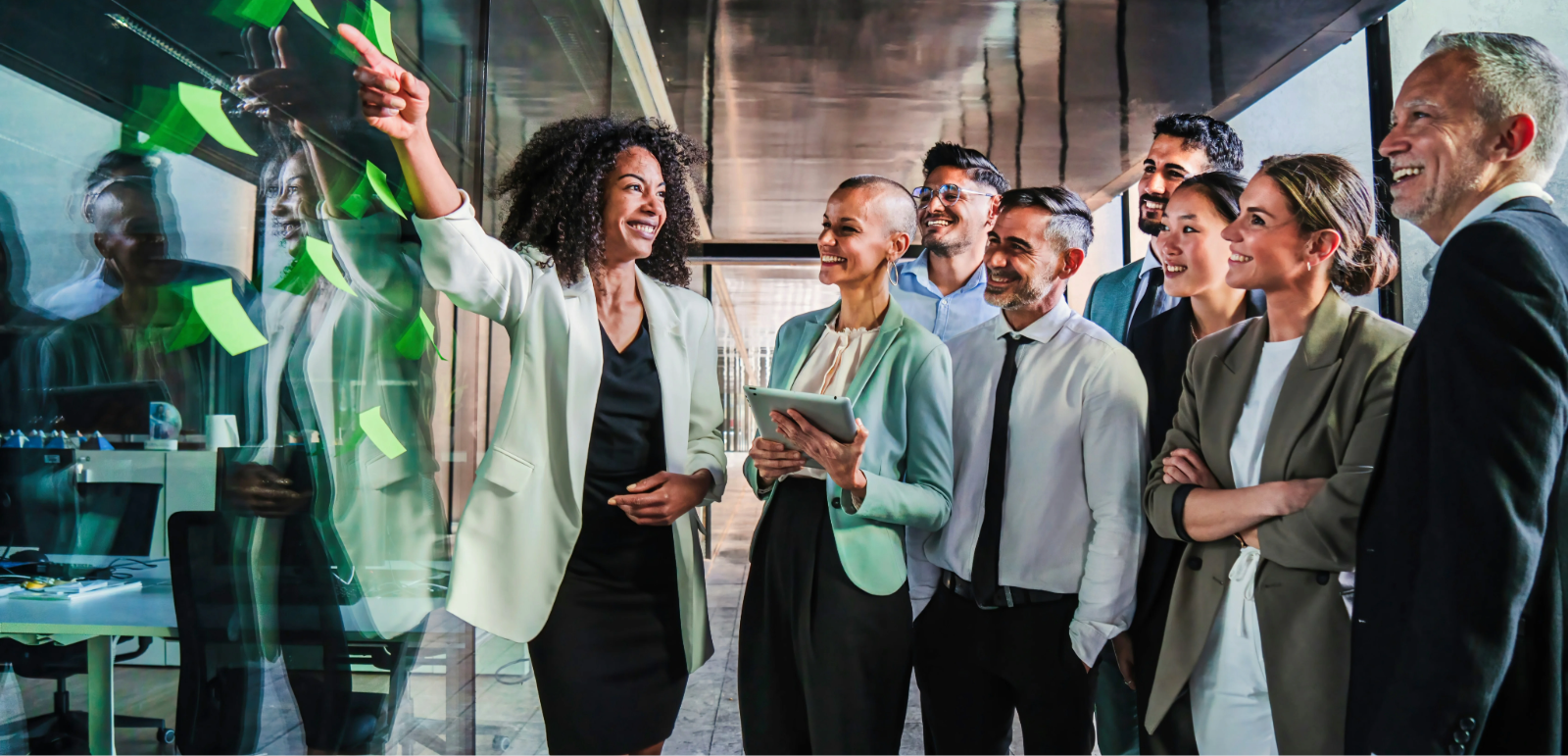 A diverse group of professionals stands discussing by a glass wall with sticky notes, led by a woman pointing and smiling.