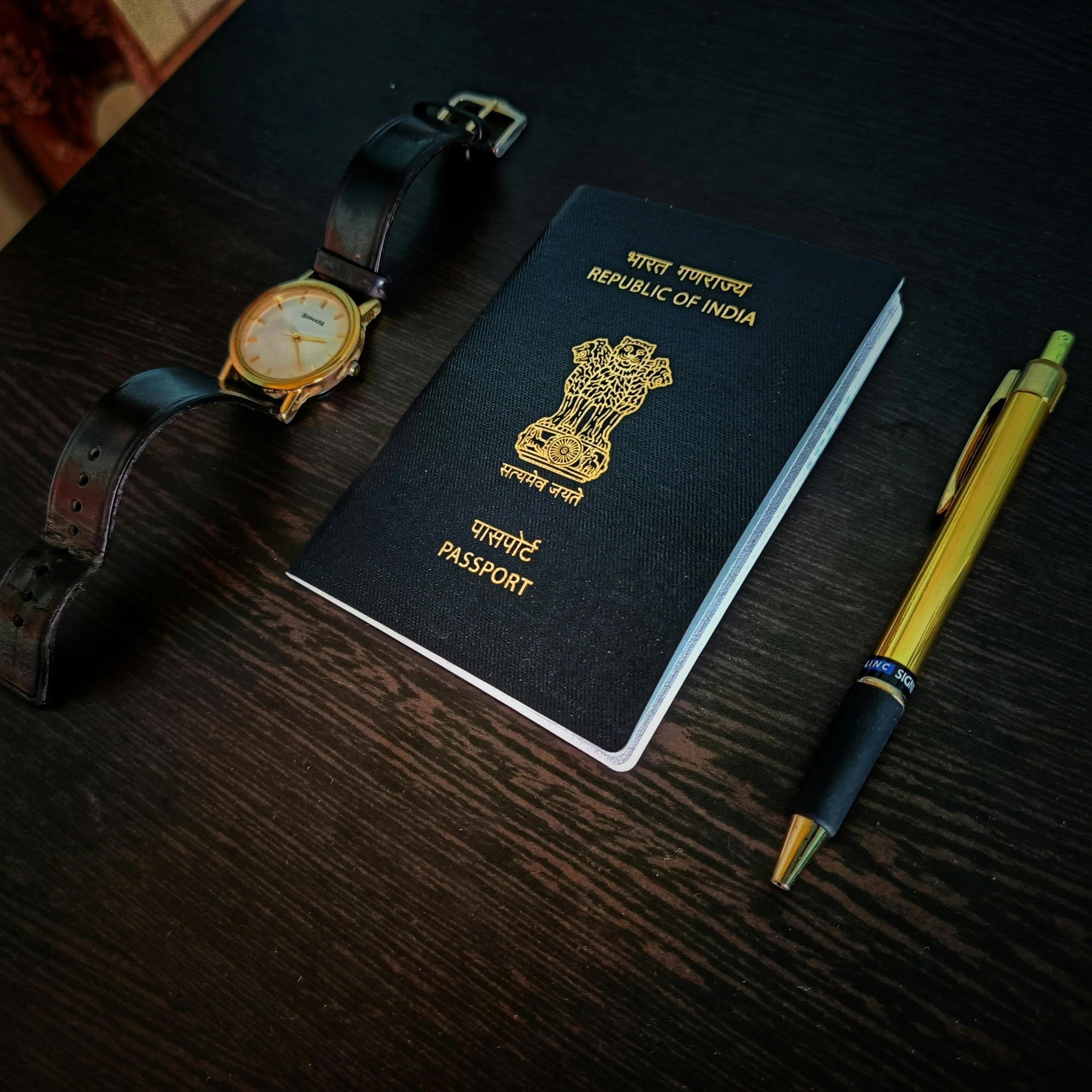 A Republic of India passport on a dark surface with a gold watch and a gold-accented pen.