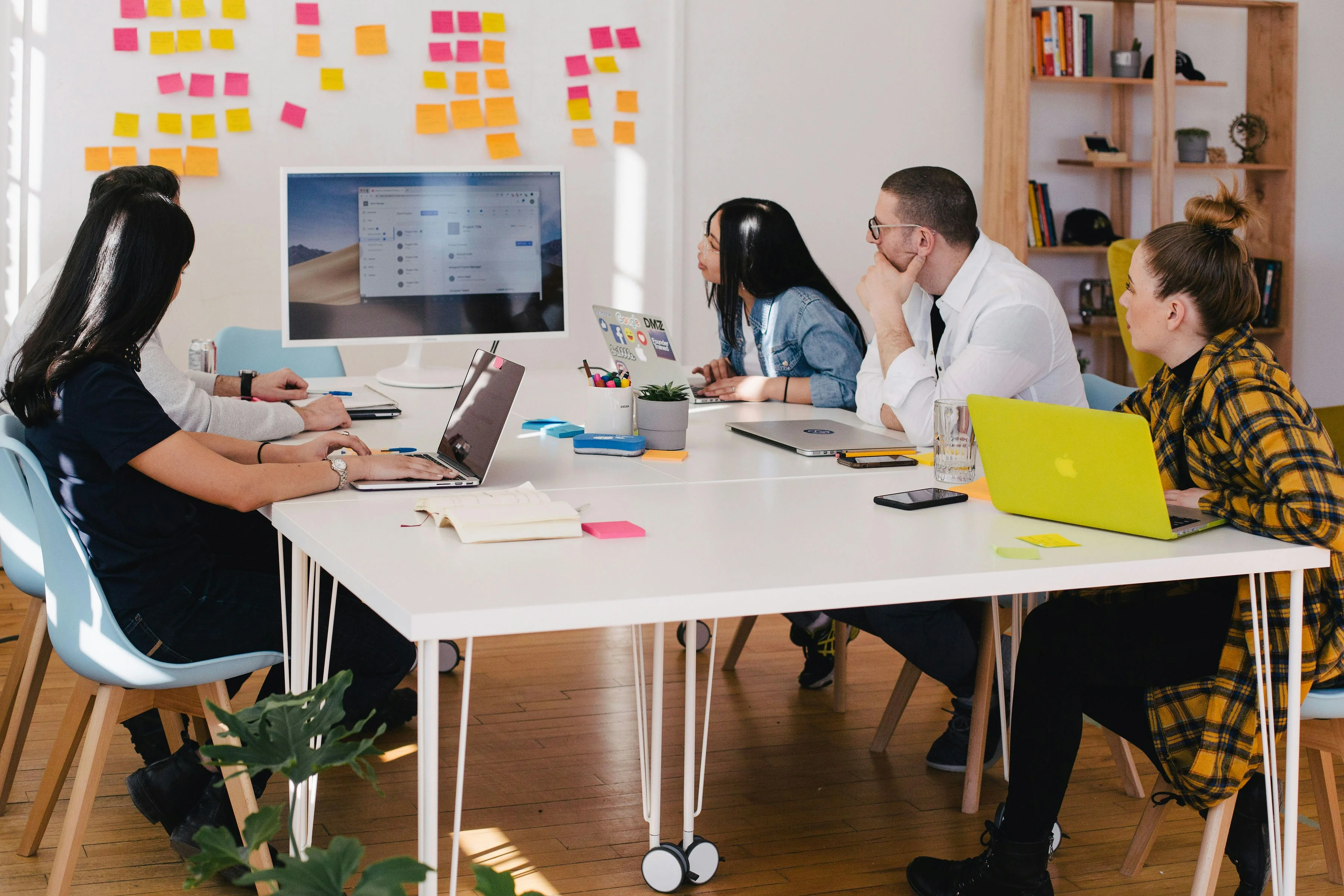 Five people in a meeting room, focused on a presentation screen. Laptops and colorful sticky notes are on the table and wall.