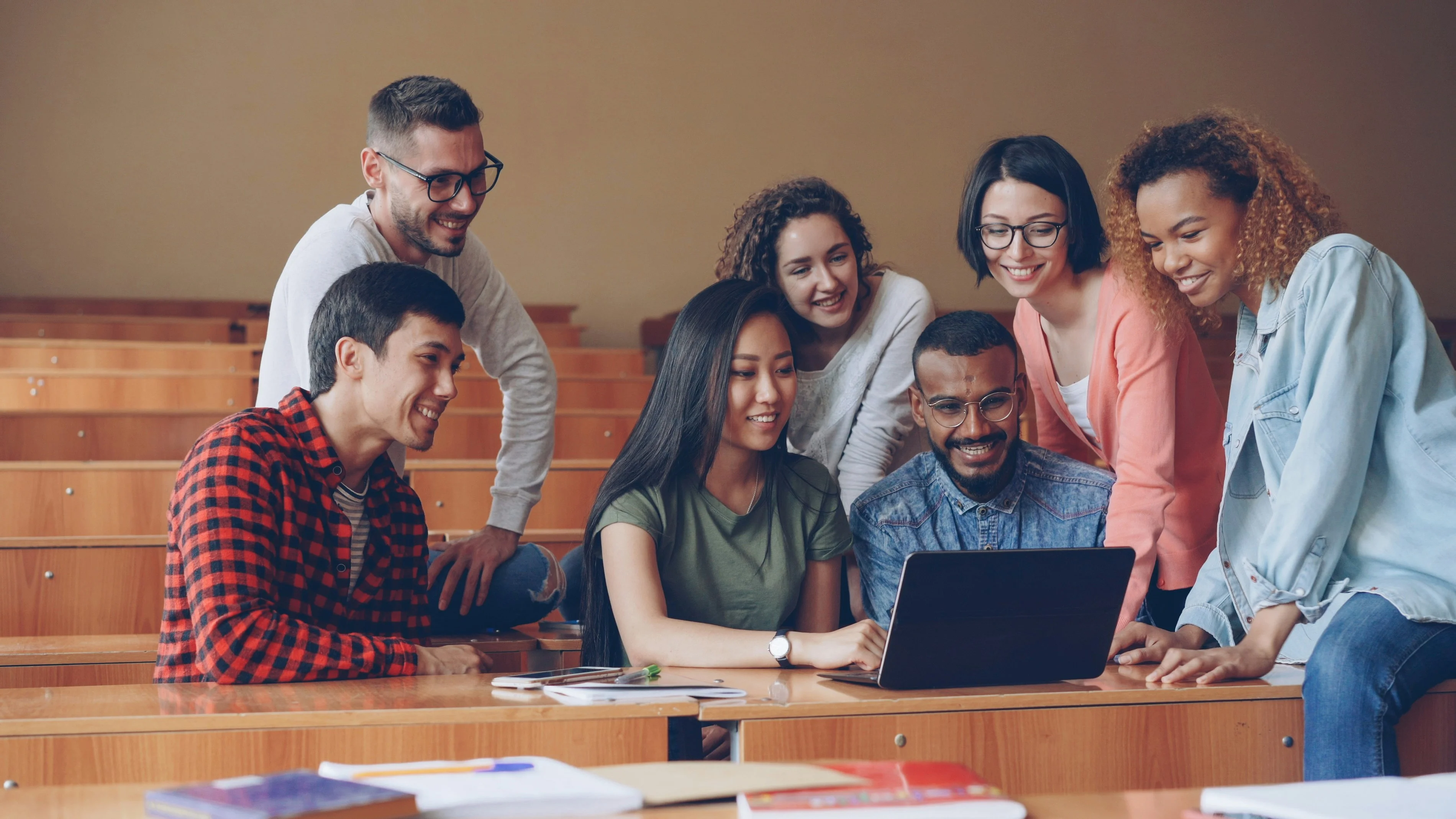 A diverse group of students gather around a laptop in a lecture hall, smiling and engaged in a collaborative activity.