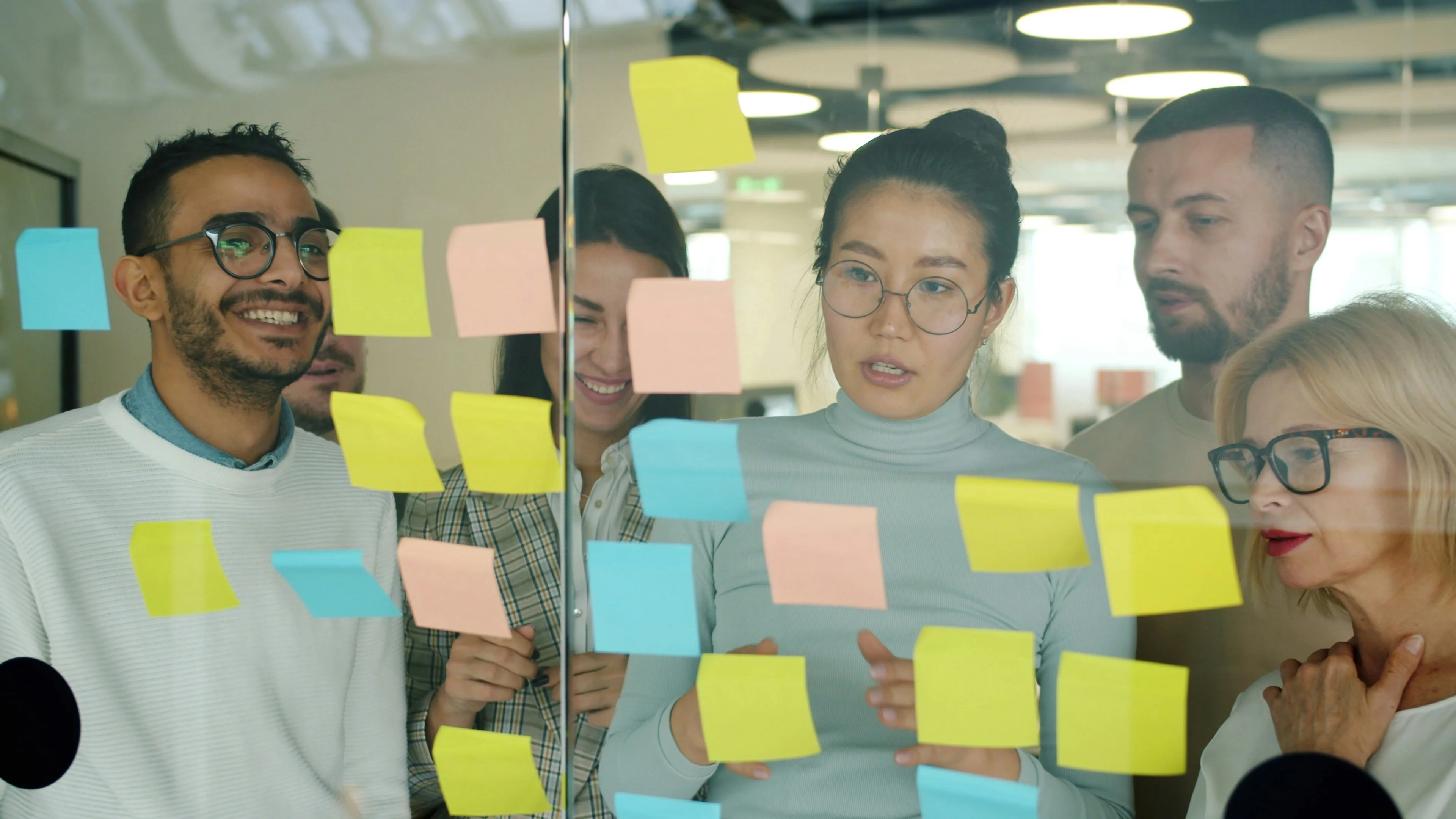 A group of five people collaborates in an office, discussing ideas and writing on colorful sticky notes attached to a glass wall.